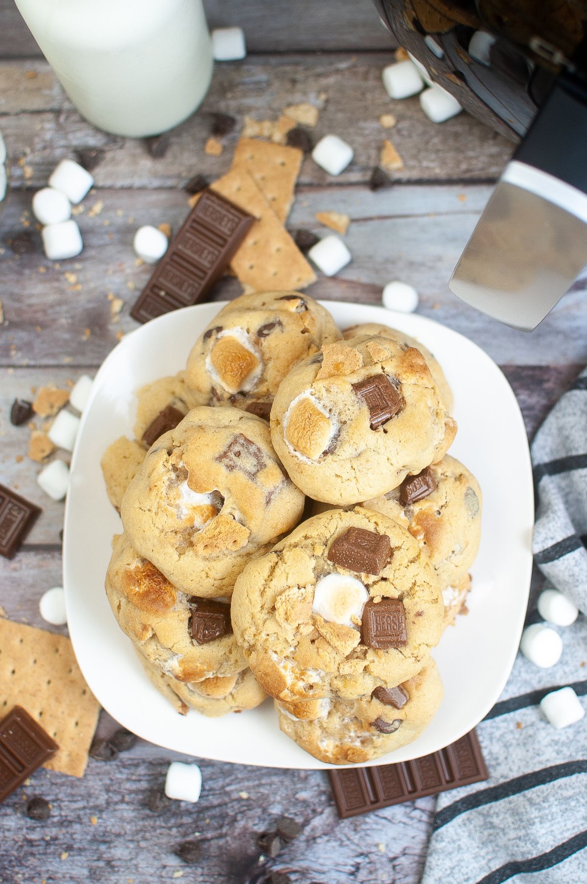 closeup view of a stack of cookies made from an Air Fryer Smores Cookie Recipe on a plate surrounded by graham crackers, chocolate bars, chocolate chips and marshmallows