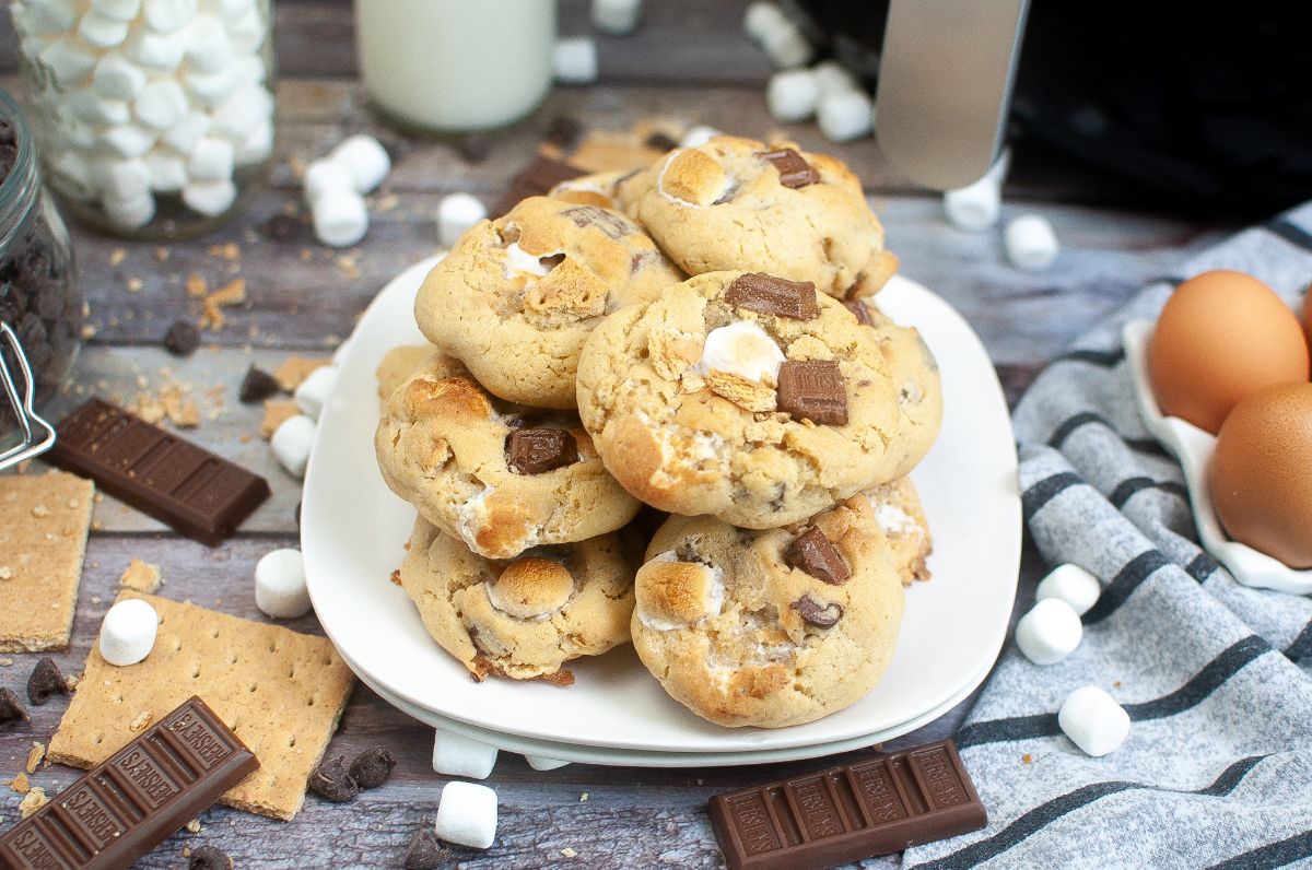 a stack of cookies made from an Air Fryer Smores Cookie Recipe on a plate surrounded by graham crackers, chocolate bars, chocolate chips and marshmallows
