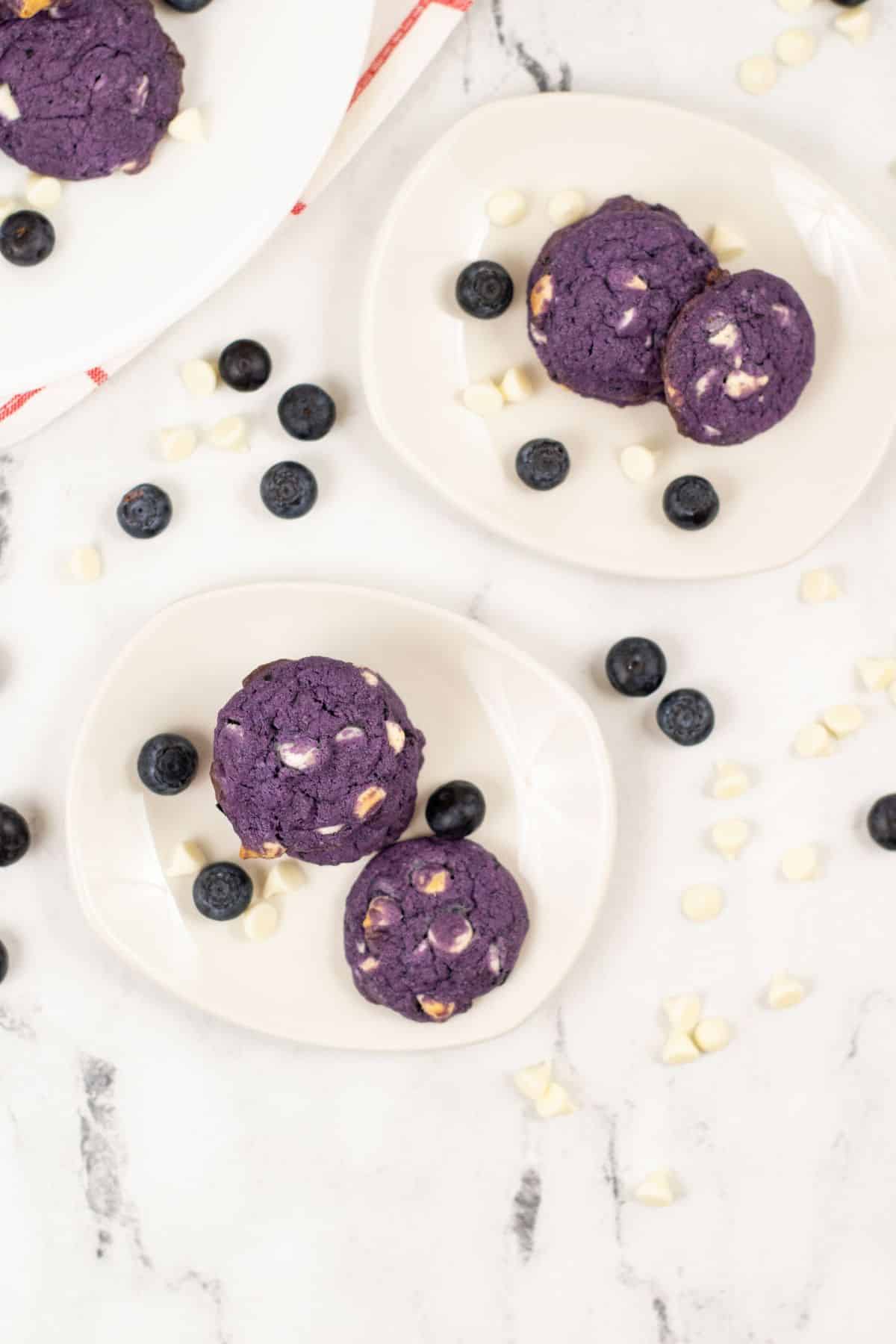 Blueberry White Chocolate Cookies on white plates surrounded by blueberries and white chocolate chips