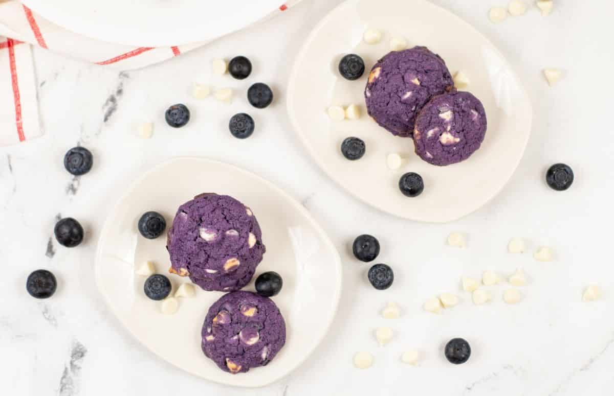 overhead view of Blueberry White Chocolate Cookies on white plates surrounded by blueberries and white chocolate chips