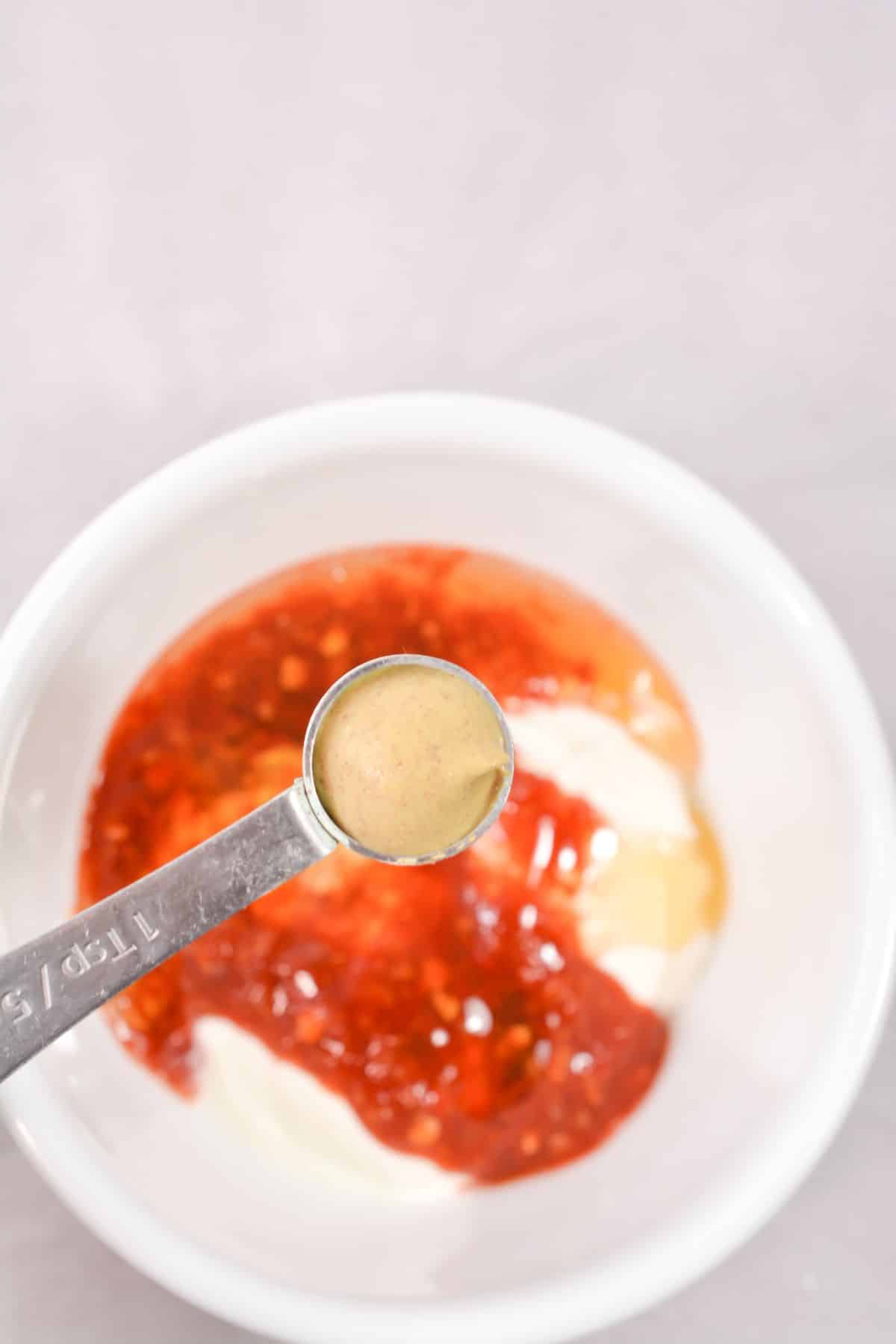 dijon mustard being added to Mayo, sour cream, chili garlic sauce, sugar-free honey in a bowl