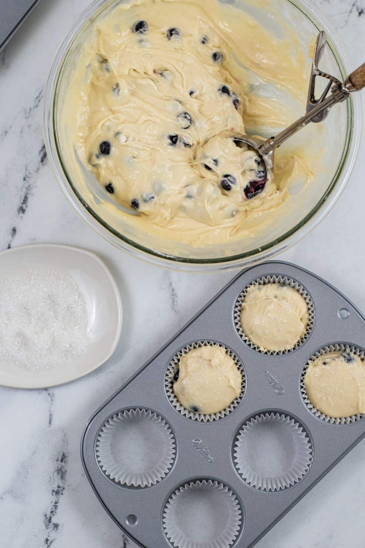 Muffin tin and muffin wrappers full with batter next to a bowl of muffin batter with a scoop in it