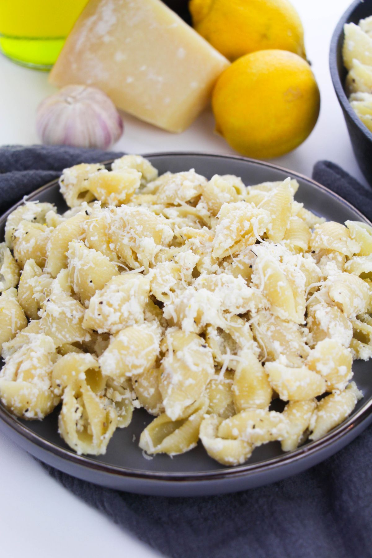 Lemon Ricotta Pasta on a plate topped with grated  parmesan cheese with more pasta in a dish in the background next to lemons, parmesan and garlic