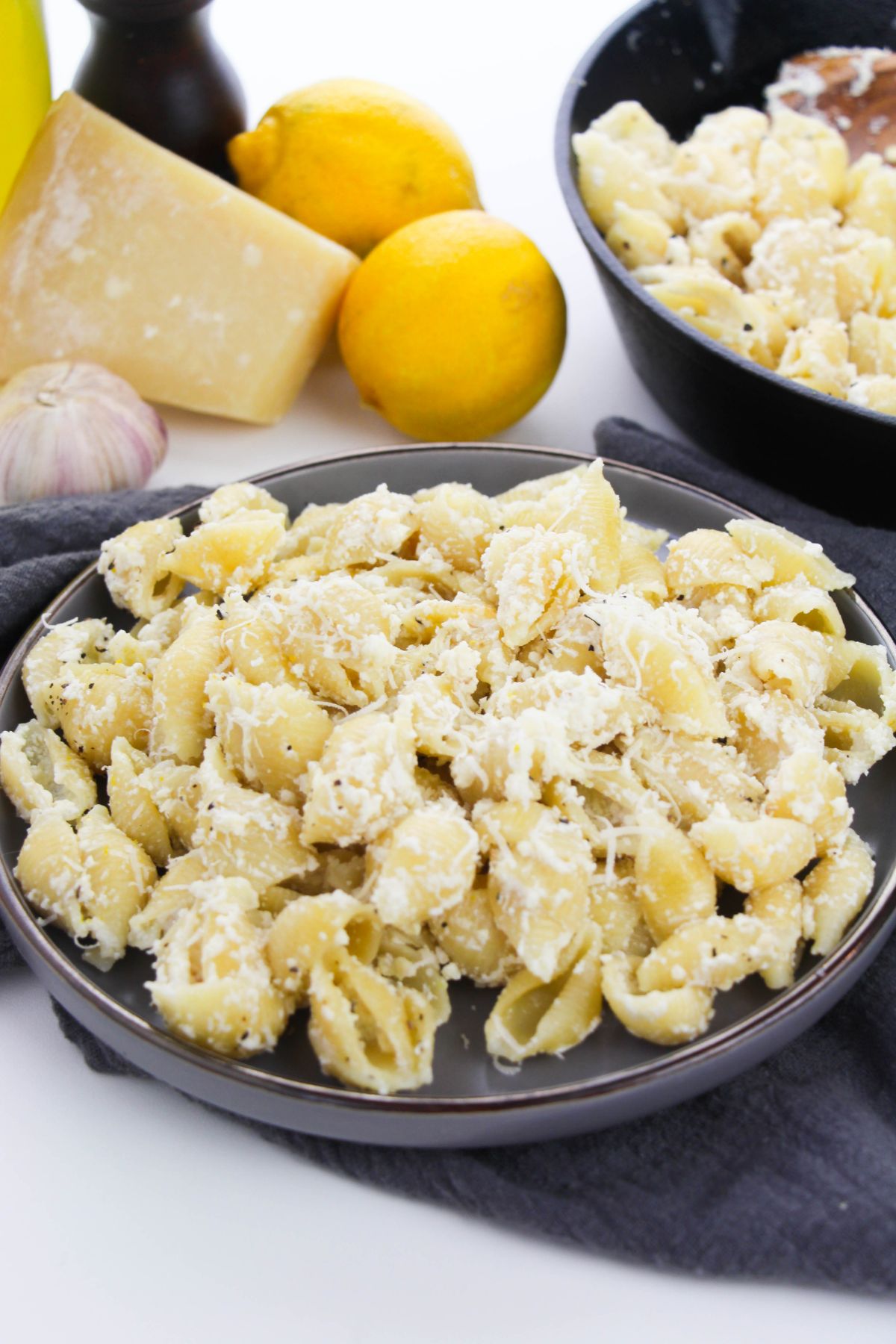 Lemon Ricotta Pasta on a plate topped with grated  parmesan cheese with more pasta in a dish in the background next to lemons, parmesan and garlic