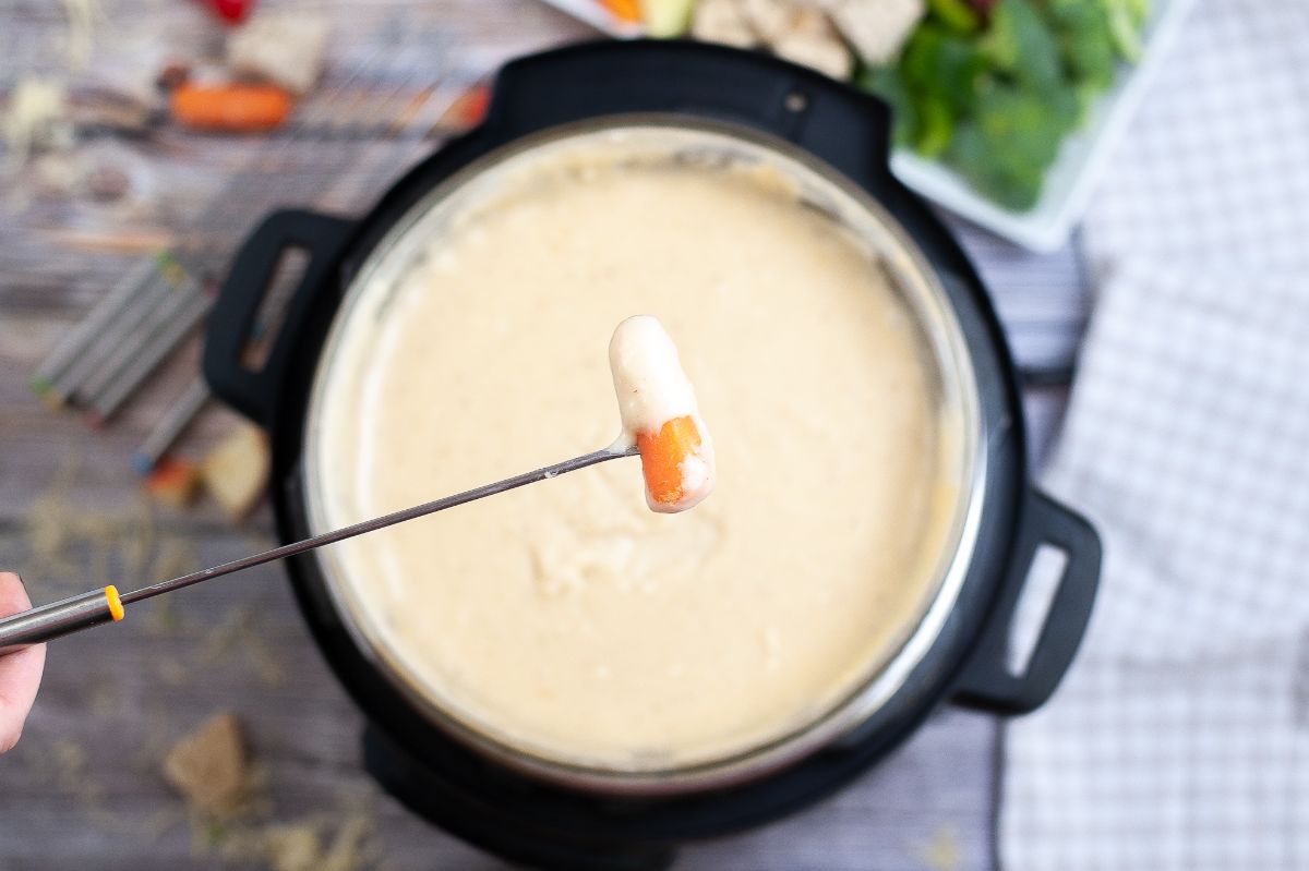 a carrot dipped in fondue on a skewer above Swiss cheese fondue made in the Instant Pot, with vegetables on the side