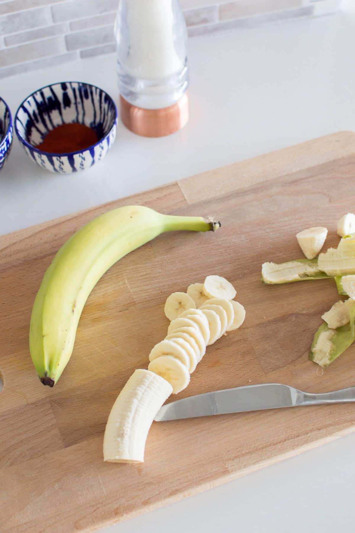 a banana next to a partially sliced banana and a banana peel on a wooden cutting board with a knife