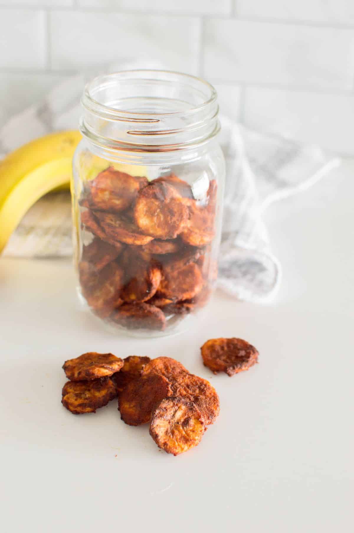 Air Fryer Banana Chips in a jar and on a table  with bananas on the side