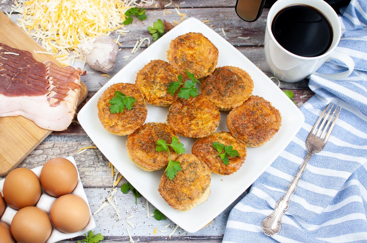 Air Fryer Egg Bites on a white plate garnished with parsley next to eggs, bacon, shredded cheese, a cup of coffee and a fork