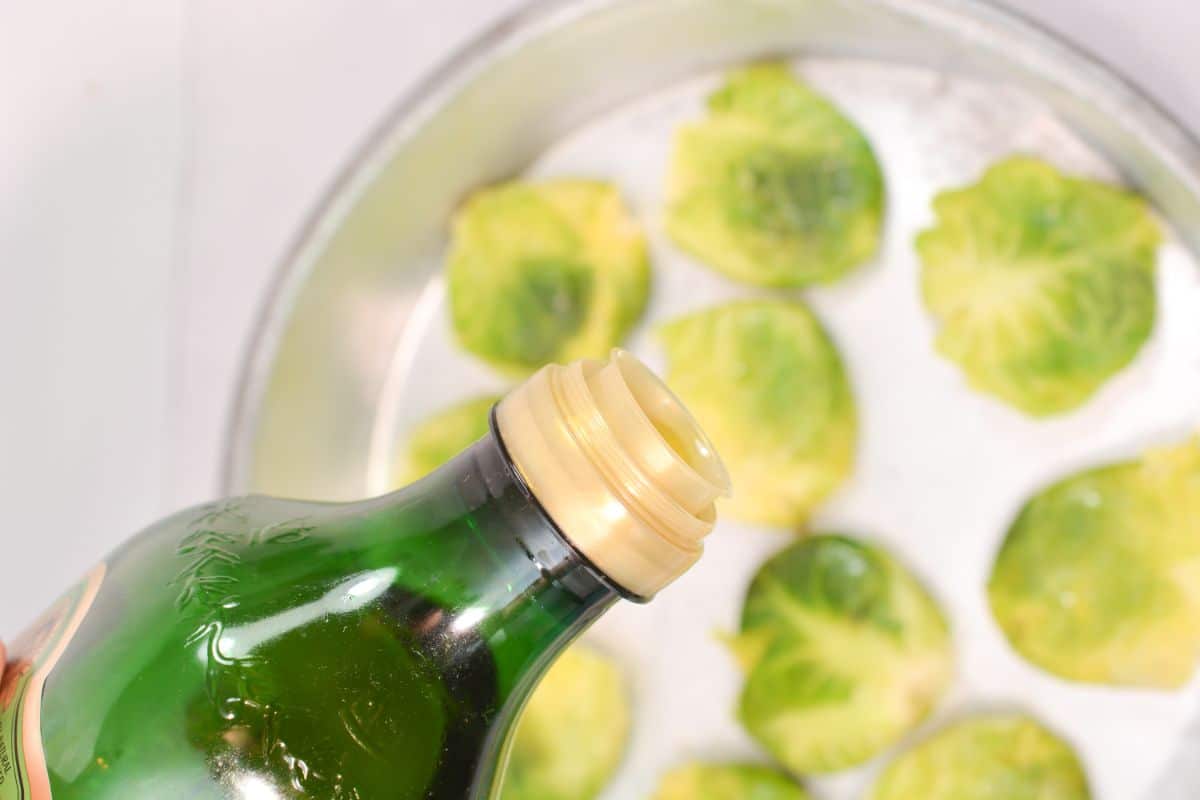 a close up of a bottle of olive oil above a blurred image of brussel sprouts in a pan