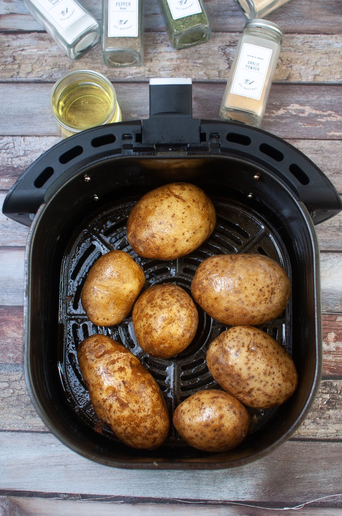 Potatoes coated with oil in the air fryer next to more oil in a jar and jars of spices