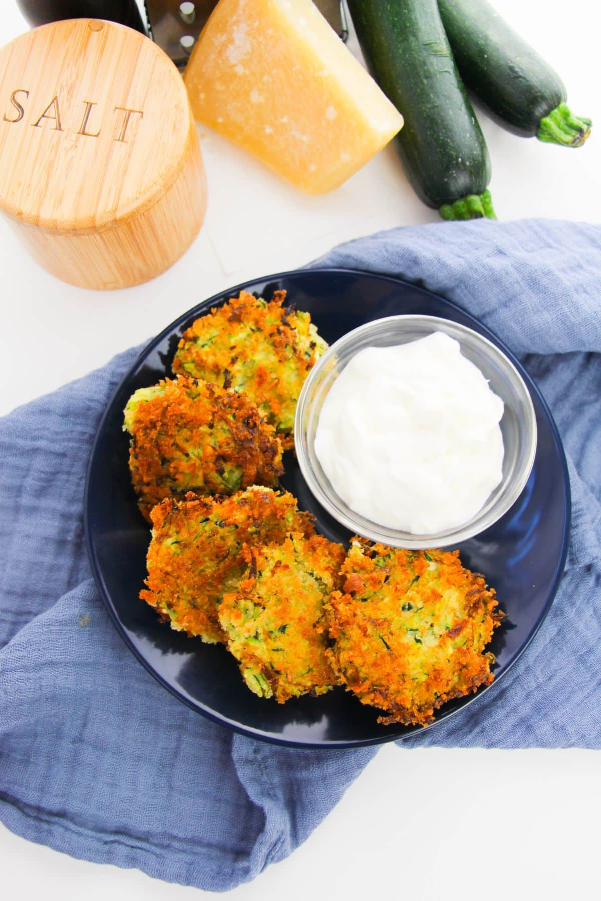 Crispy Zucchini Fritters on a plate with dipping sauce, on a blue cloth with salt, cheese and zucchini in the background