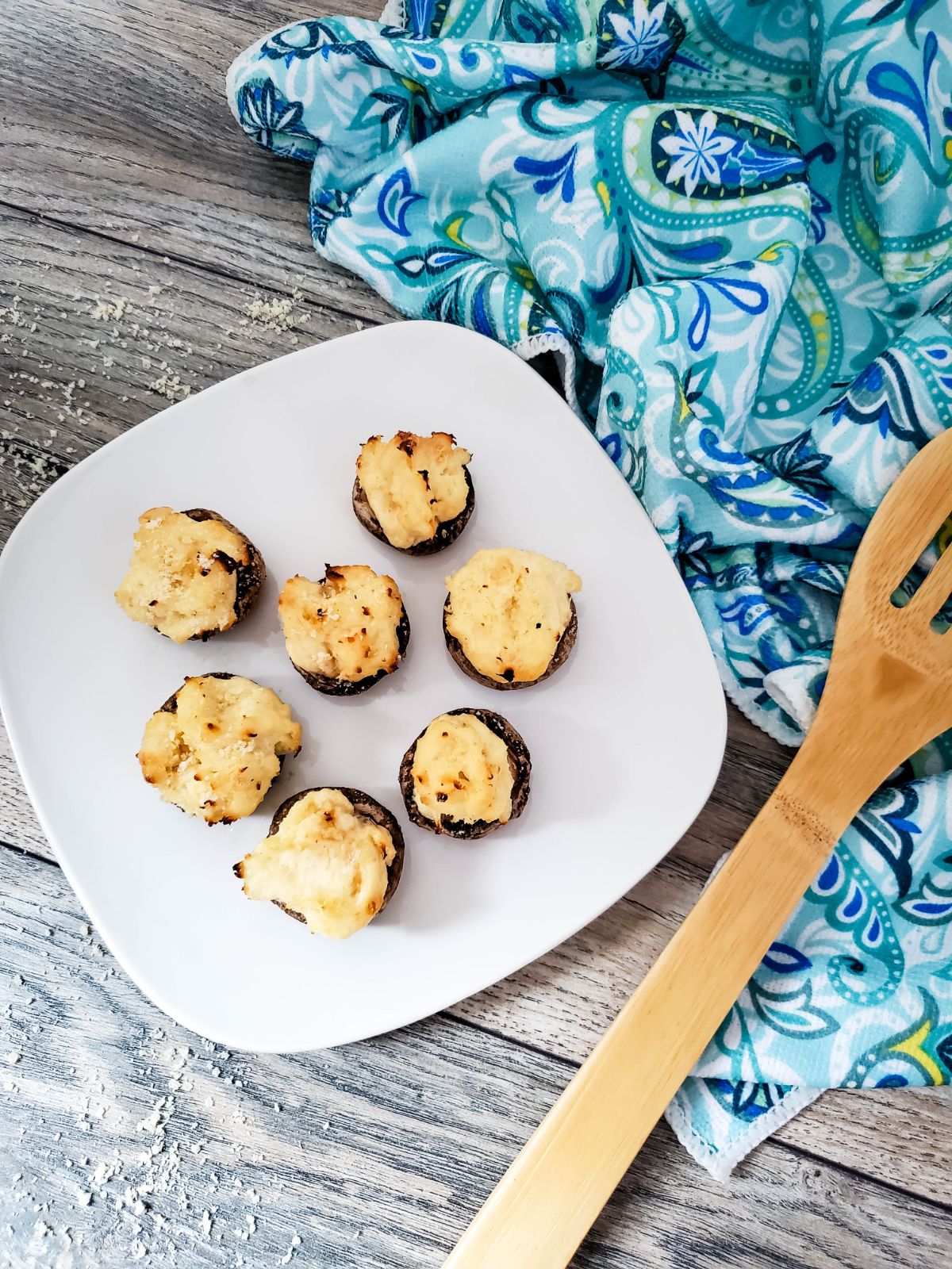 Grilled Stuffed Mushrooms with Cream Cheese on a white plate with wooden slotted spoon on the side on a blue decorative cloth