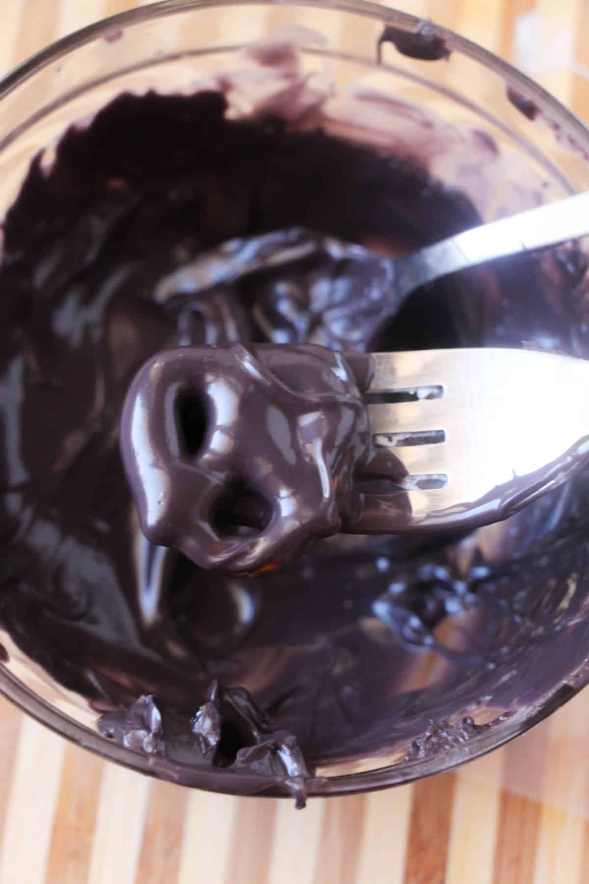 a fork holding a Pretzel coated with melted black candy above the rest of the melted candy in a glass bowl