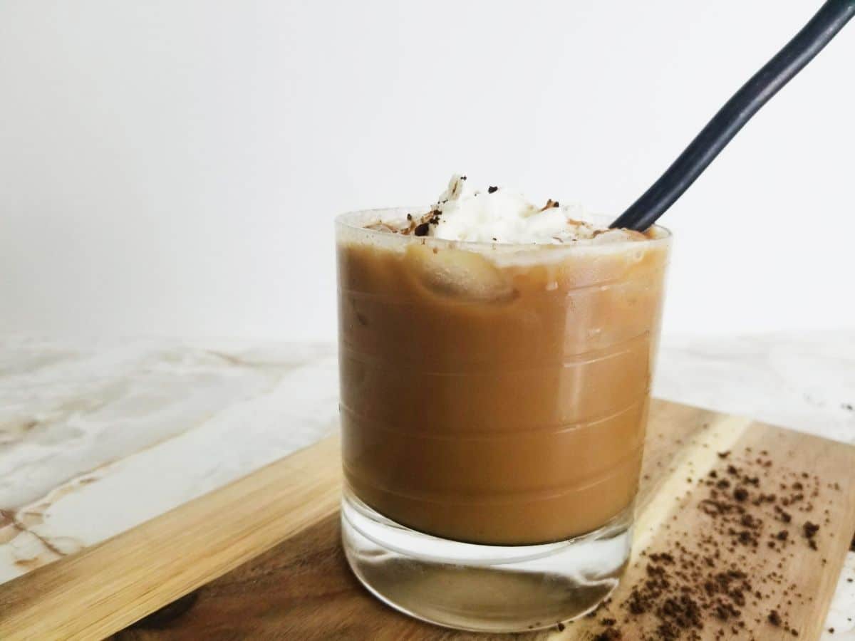 Iced Coffee topped with whipped cream and cinnamon in a glass with a straw in it, on a wooden board.