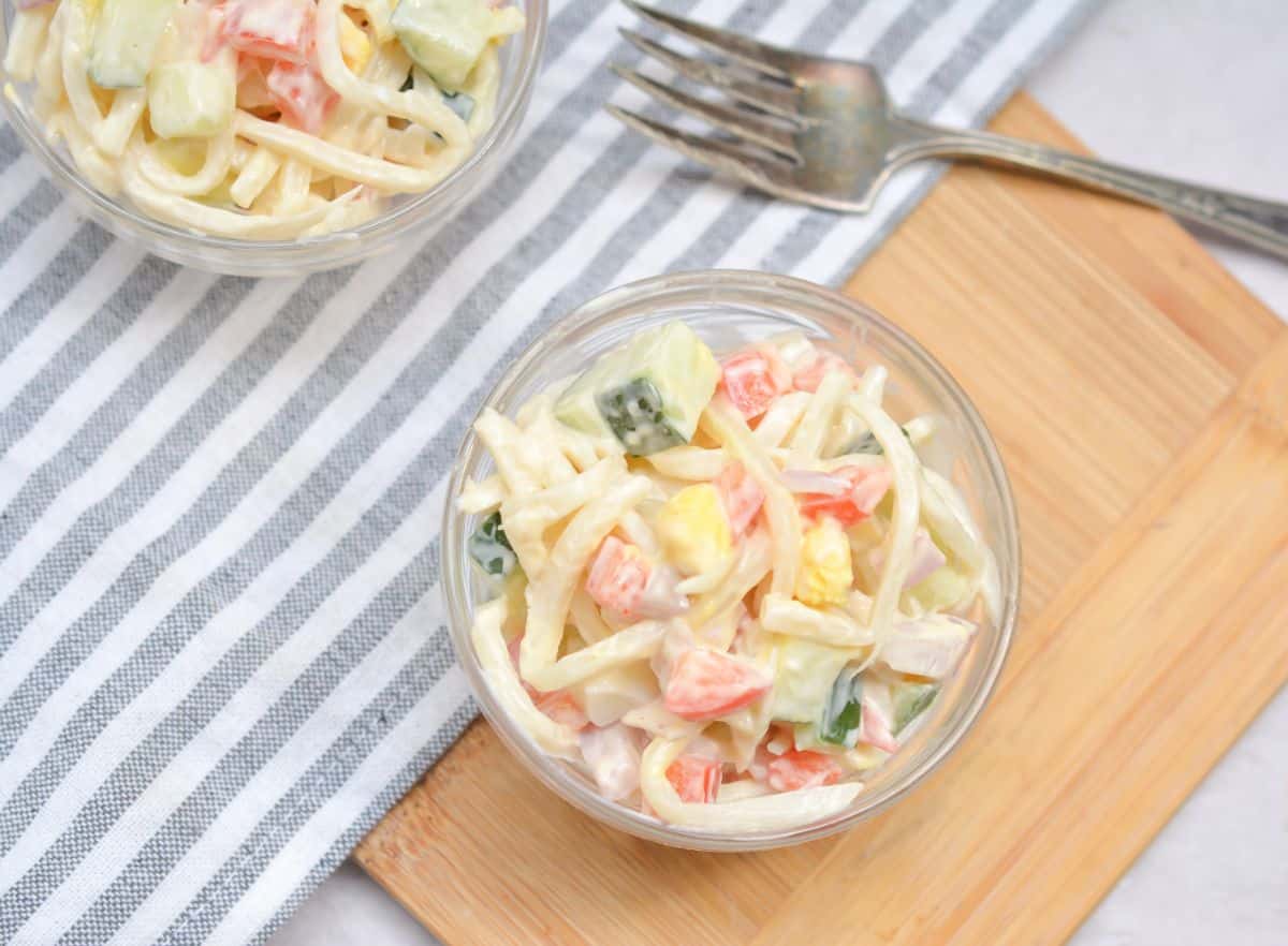 Keto Pasta Salad in a glass bowl on a wood board next to a fork with another bowl of salad behind it on a gray and white striped cloth