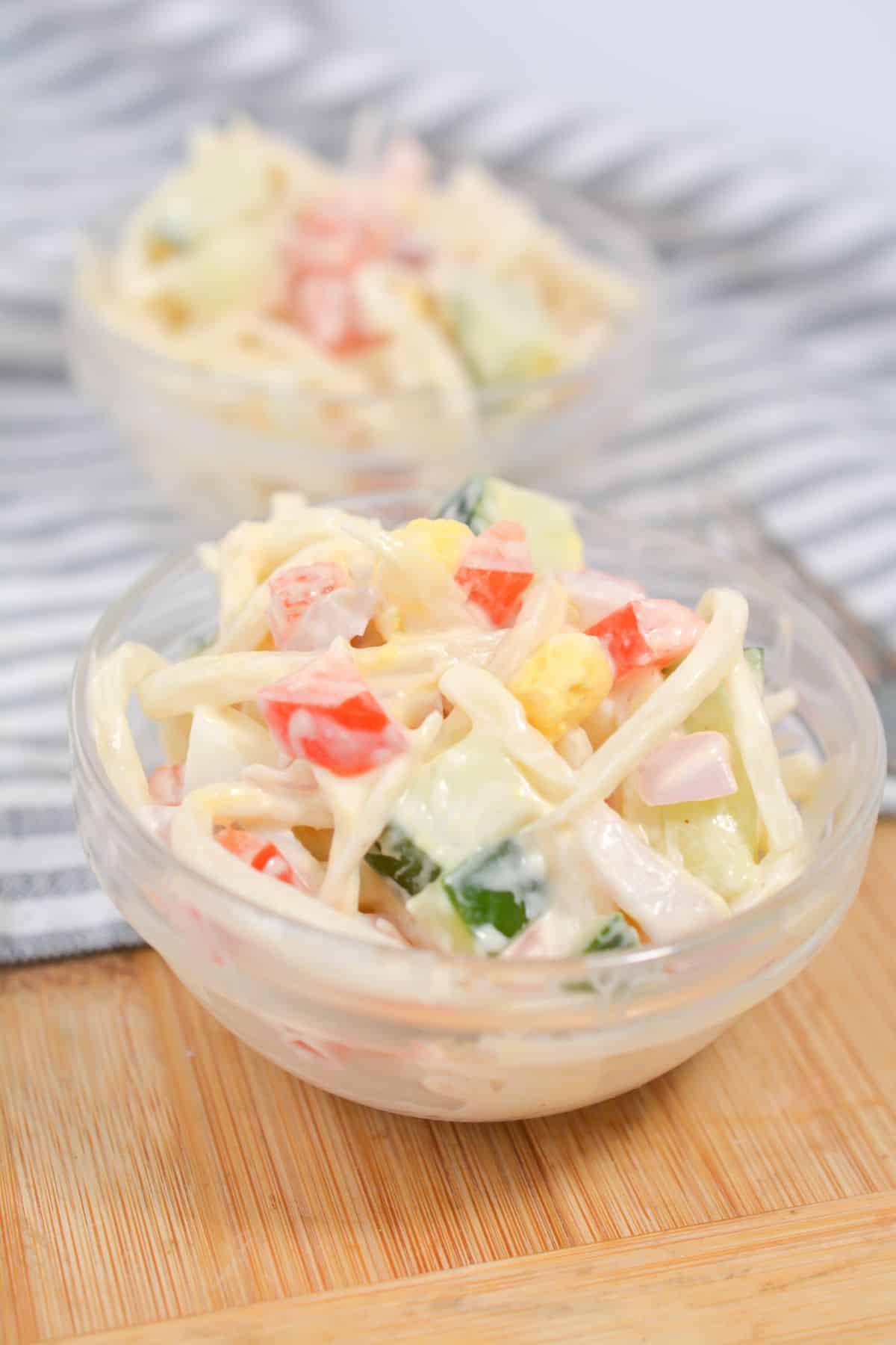 Keto Pasta Salad in a glass bowl on a wood board with another bowl of salad blurred behind it on a gray and white striped cloth