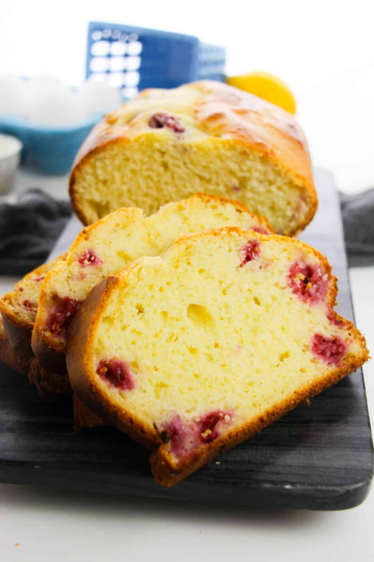 sliced Lemon Raspberry Loaf on a wooden plate with flour, lemon, and egg on the side