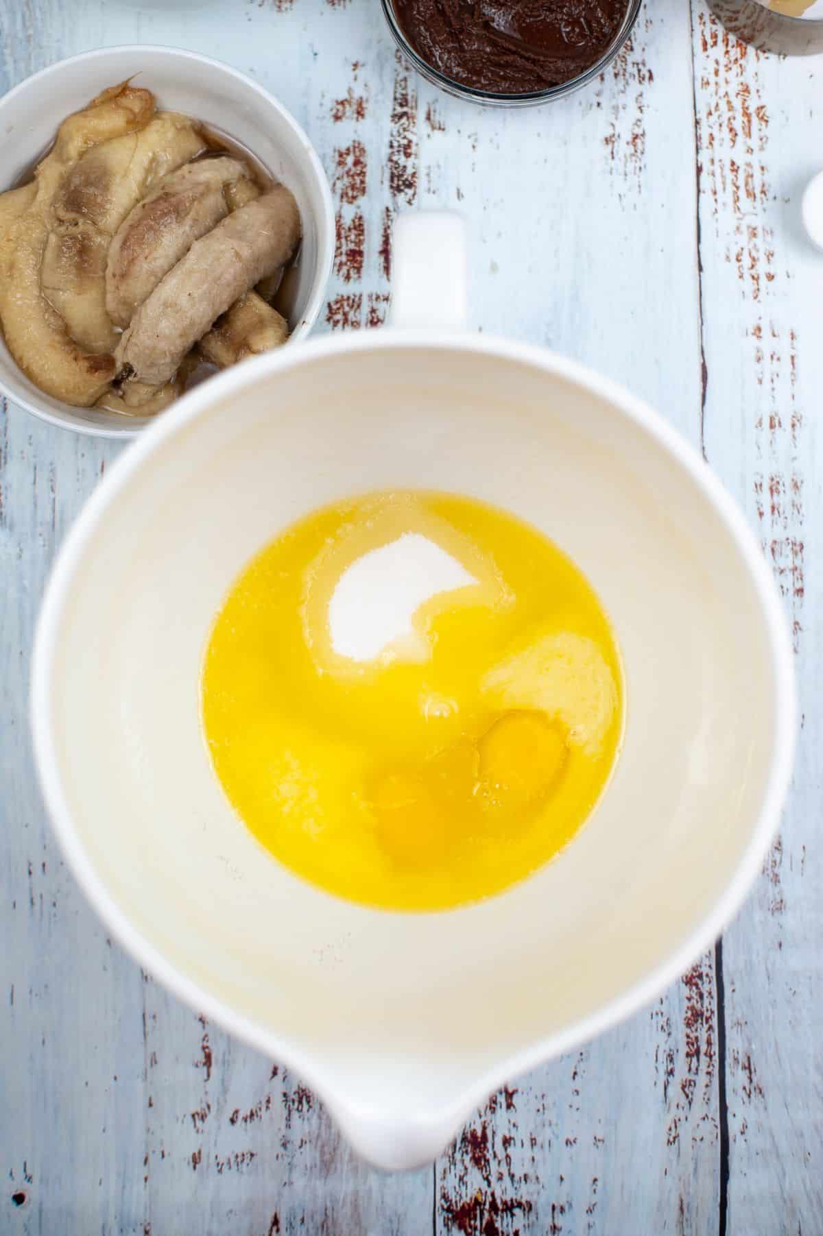 Melted butter, eggs, and sugar in a large mixing bowl next to a bowl of bananas