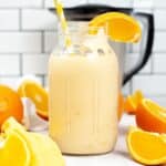 A glass jar filled with an orange milkshake, topped with a yellow striped straw and an orange slice, surrounded by more orange slices and a blender in the background.