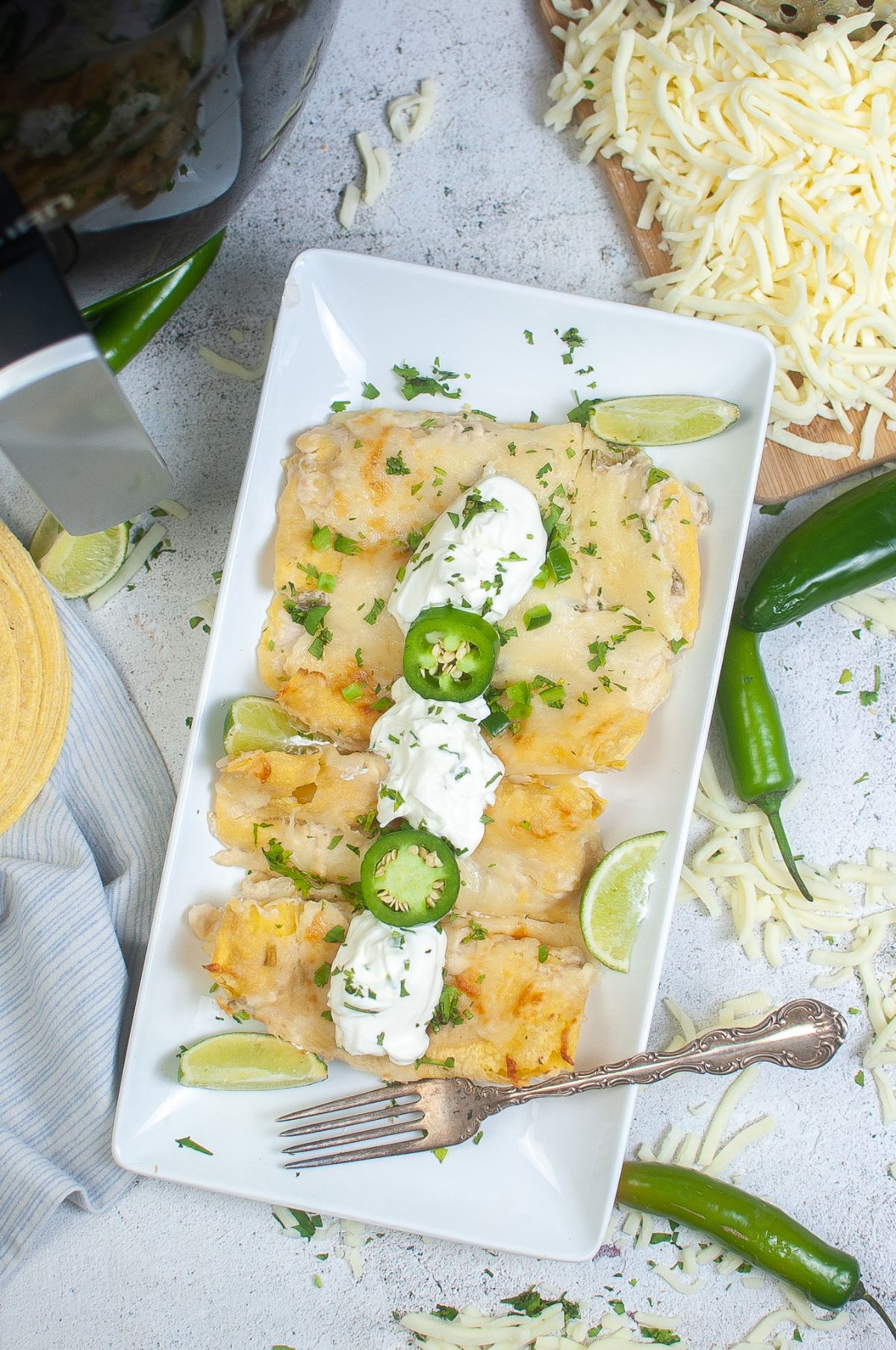 overhead view of Air Fryer Cheese Enchiladas on a plate garnished with sour cream, jalapenos, green onions, and cilantro, next to a fork with shredded cheese, jalapenos and an air fryer in the background