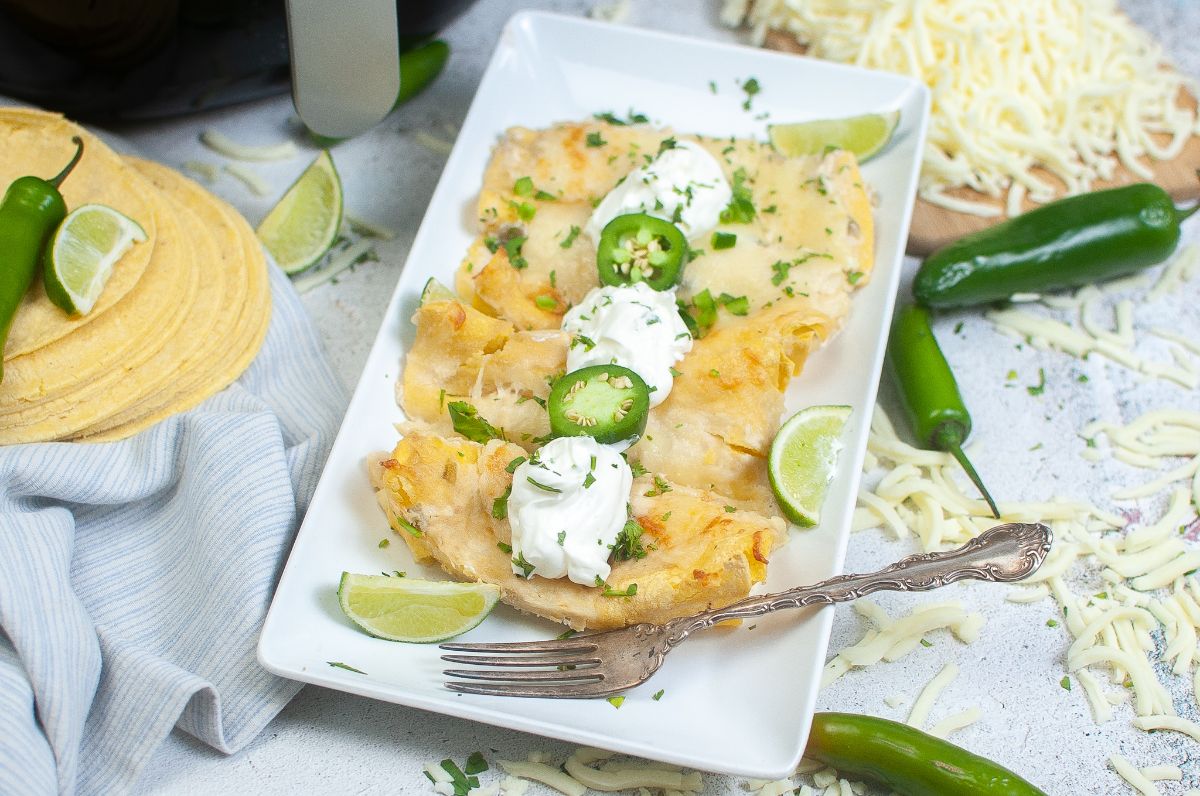 Air Fryer Cheese Enchiladas on a plate garnished with sour cream, jalapenos, green onions, and cilantro, next to a fork with shredded cheese, jalapenos and an air fryer in the background