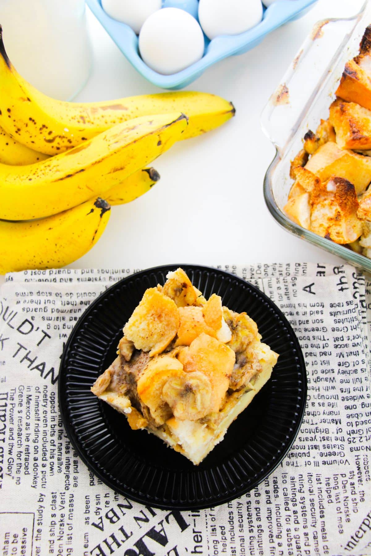 Banana Bread Pudding on a plate with bananas and eggs on the side.