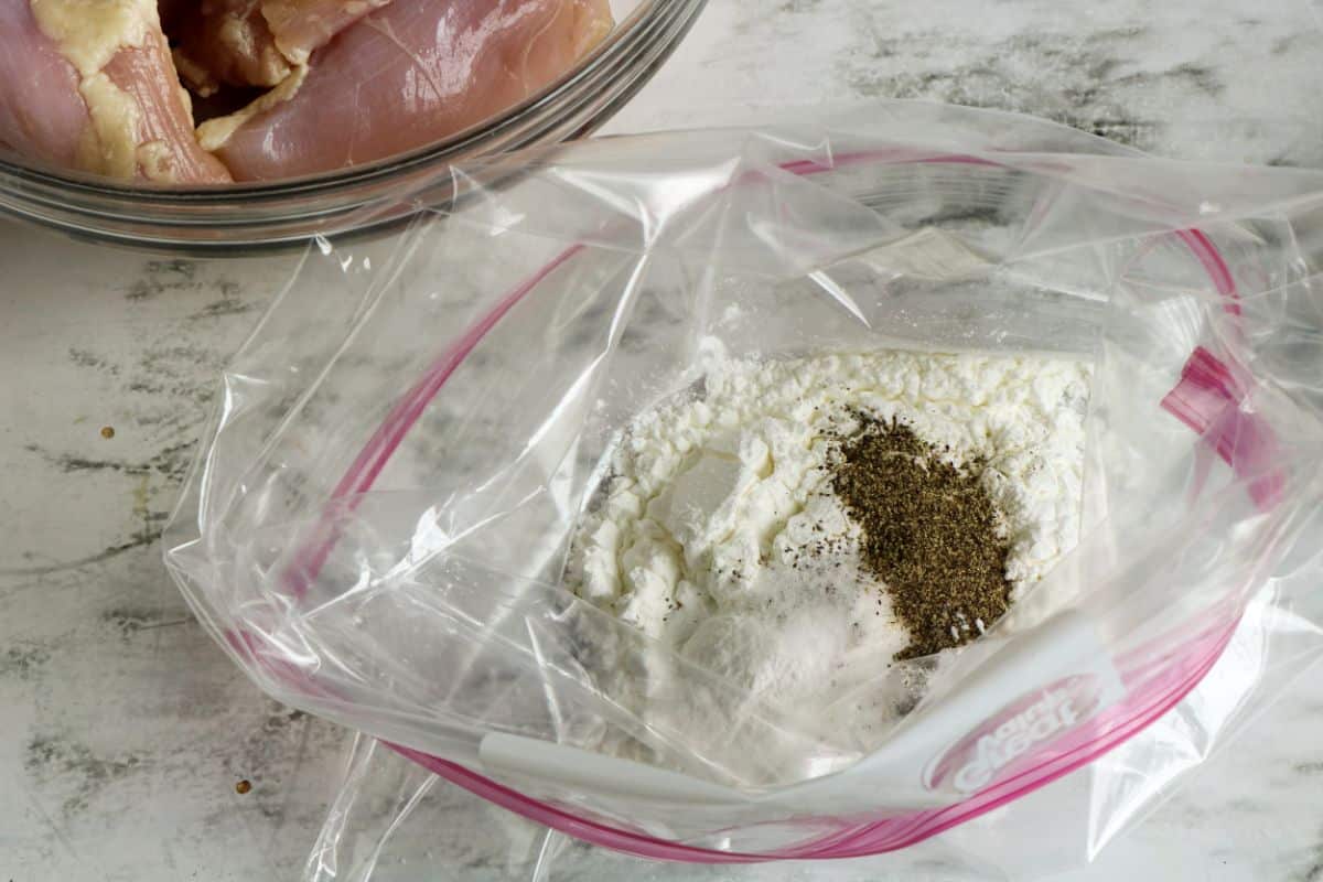 Cornstarch, salt, and pepper in a gallon-sized freezer bag next to raw chicken in a glass bowl