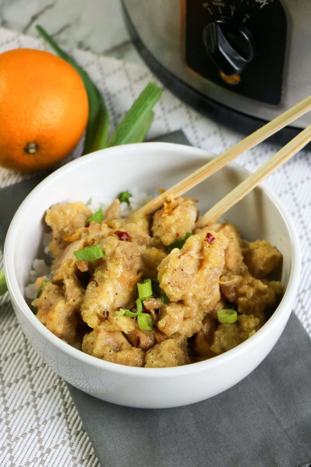 Crockpot Orange Chicken and wooden chopsticks in a white bowl with an orange, green onions, and a crockpot in the background