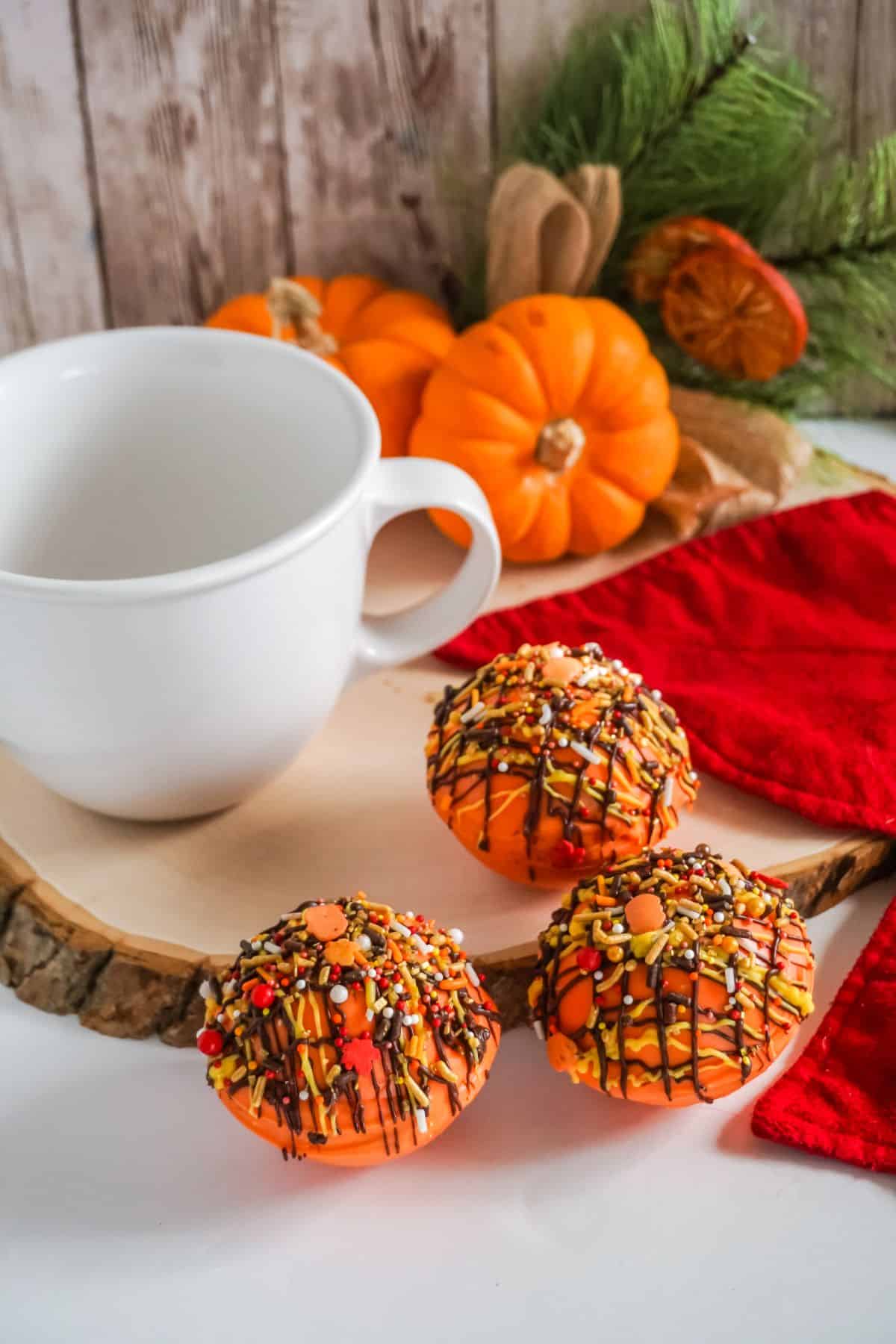 Pumpkin Spice Hot Cocoa Bombs on a rustic tree chopping board with pumpkin and white mug on the side.