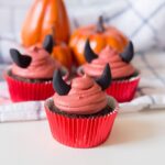 Three Halloween Devil Cupcakes with red frosting and black fondant horns in red wrappers are displayed in front of small pumpkins on a checkered cloth.