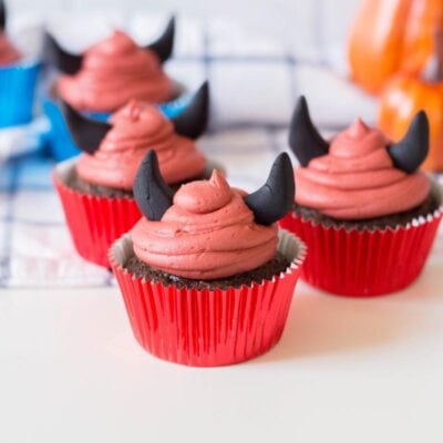 Three Halloween Devil Cupcakes with red frosting and black fondant horns sit on a white surface in front of small pumpkins. Two more similarly decorated cupcakes are in the background.