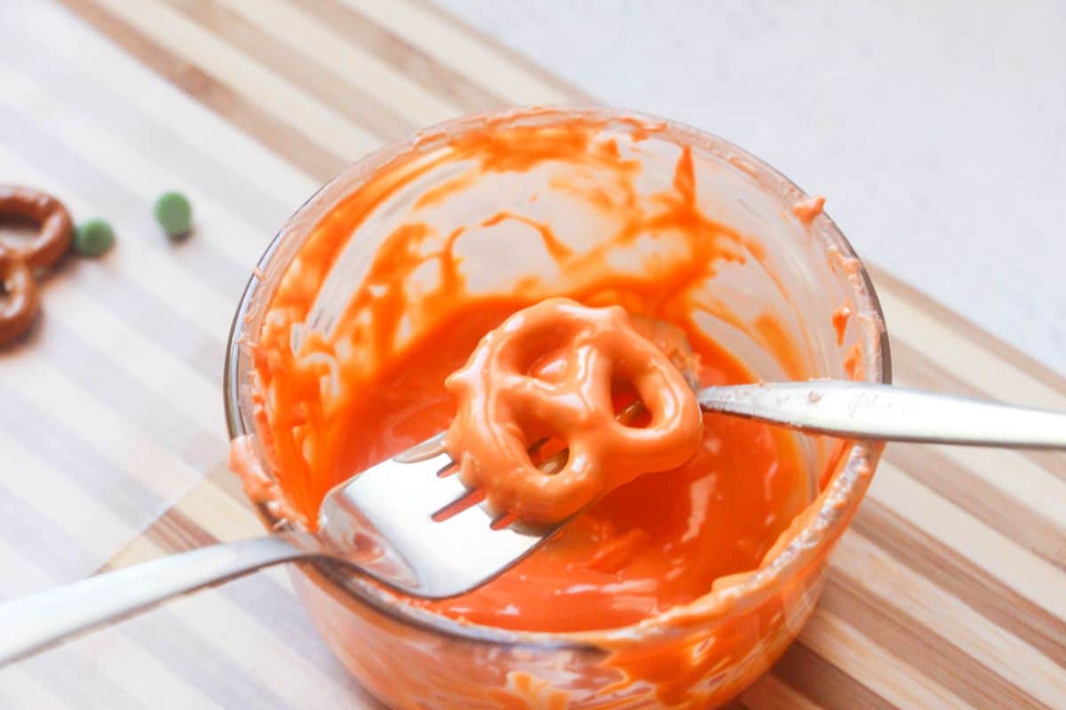 a fork holding a Pretzel coated with orange melted candy above a glass bowl of melted orange candy coating