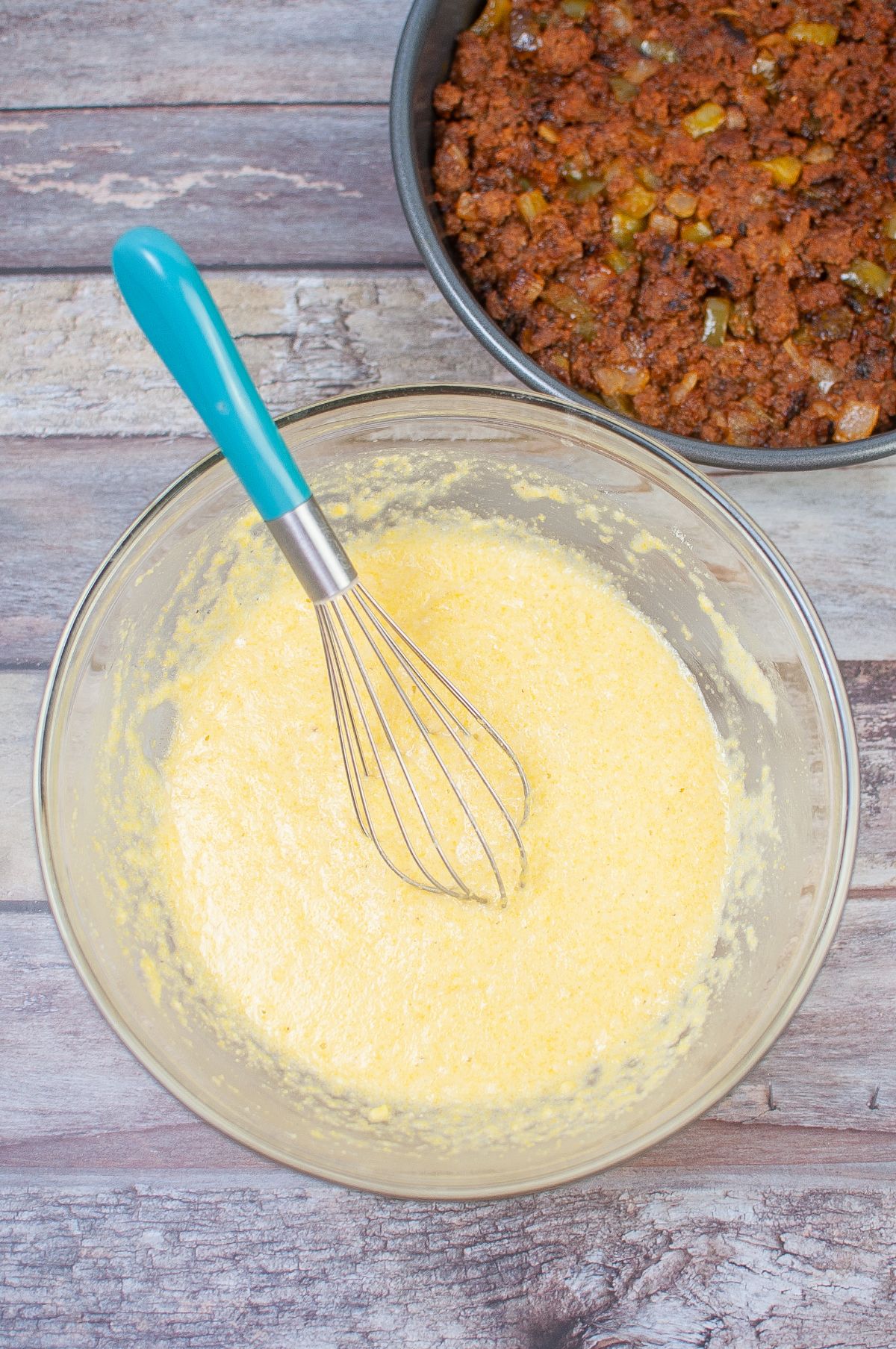 Cornbread batter in a mixing bowl with whisker next to the meat mixture in a pie pan.