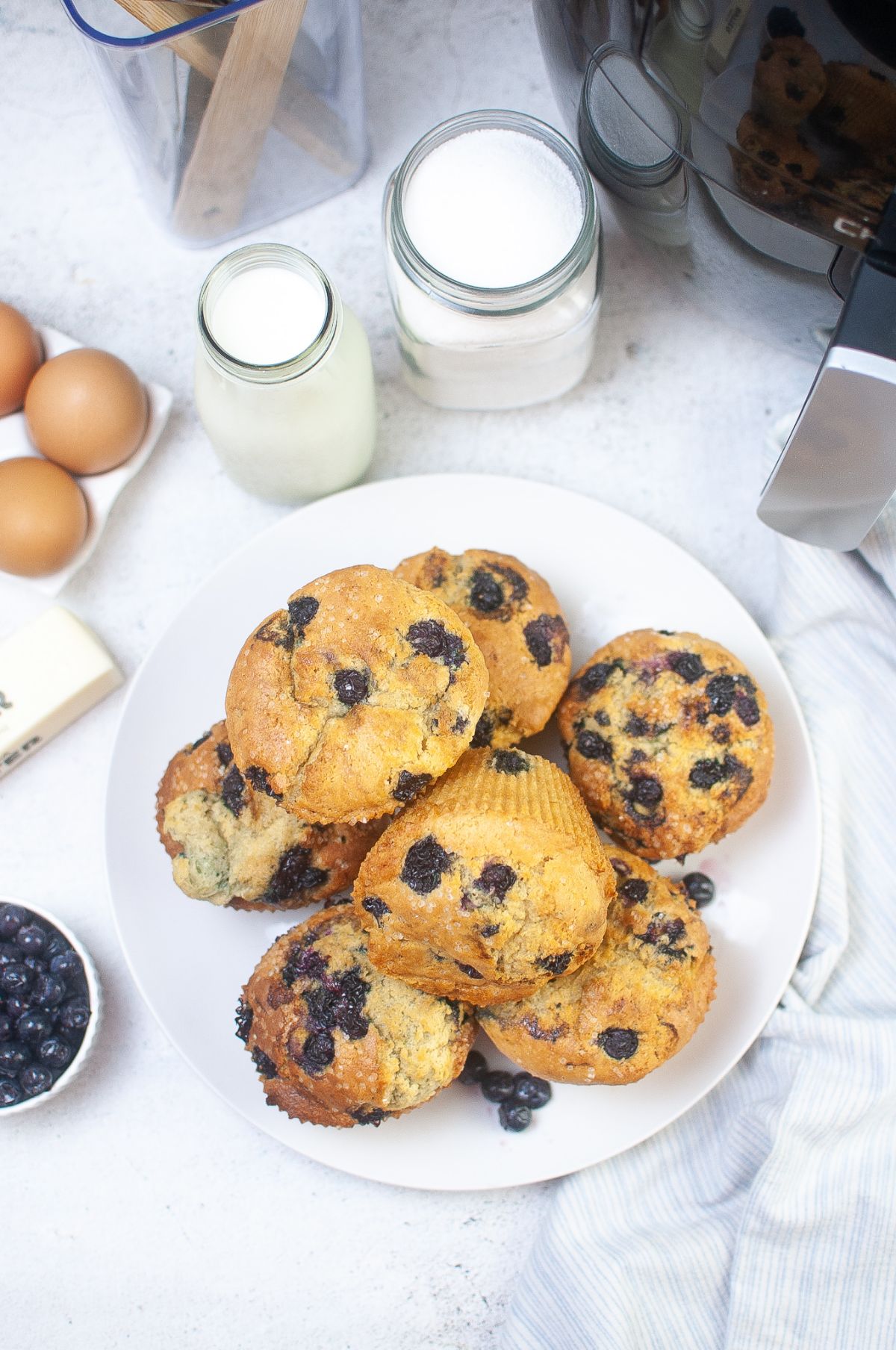 Air Fryer Blueberry Muffins on a serving plate with blueberries, air fryer and other ingredients on the side.