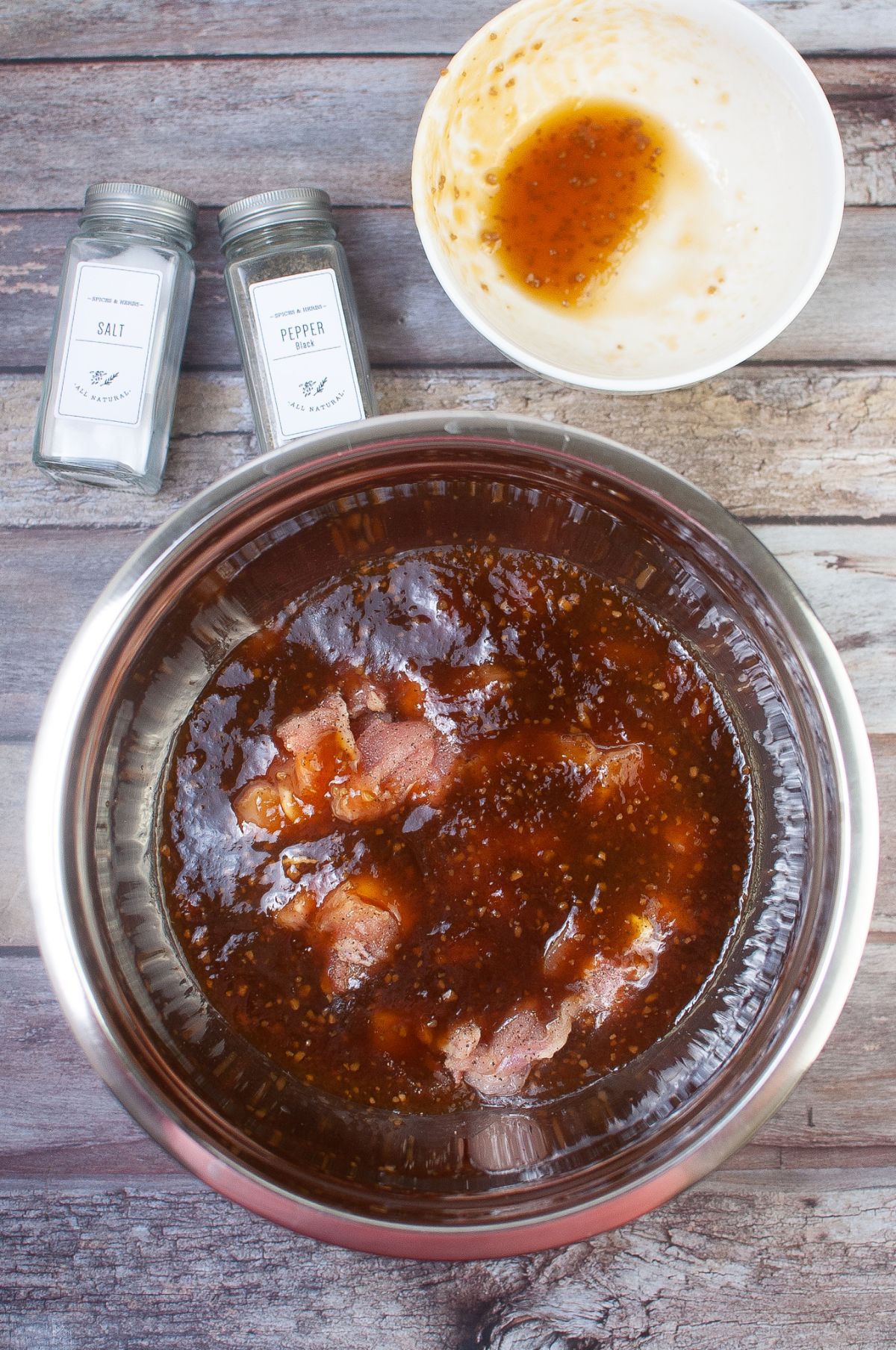 Marinade and chicken in a large bowl with pepper and salt on the side.