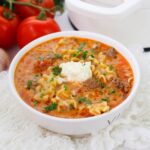 A bowl of One Pot Lasagna Soup with ground meat, diced tomatoes, pasta, and a dollop of ricotta cheese, garnished with chopped parsley. Fresh tomatoes are in the background.