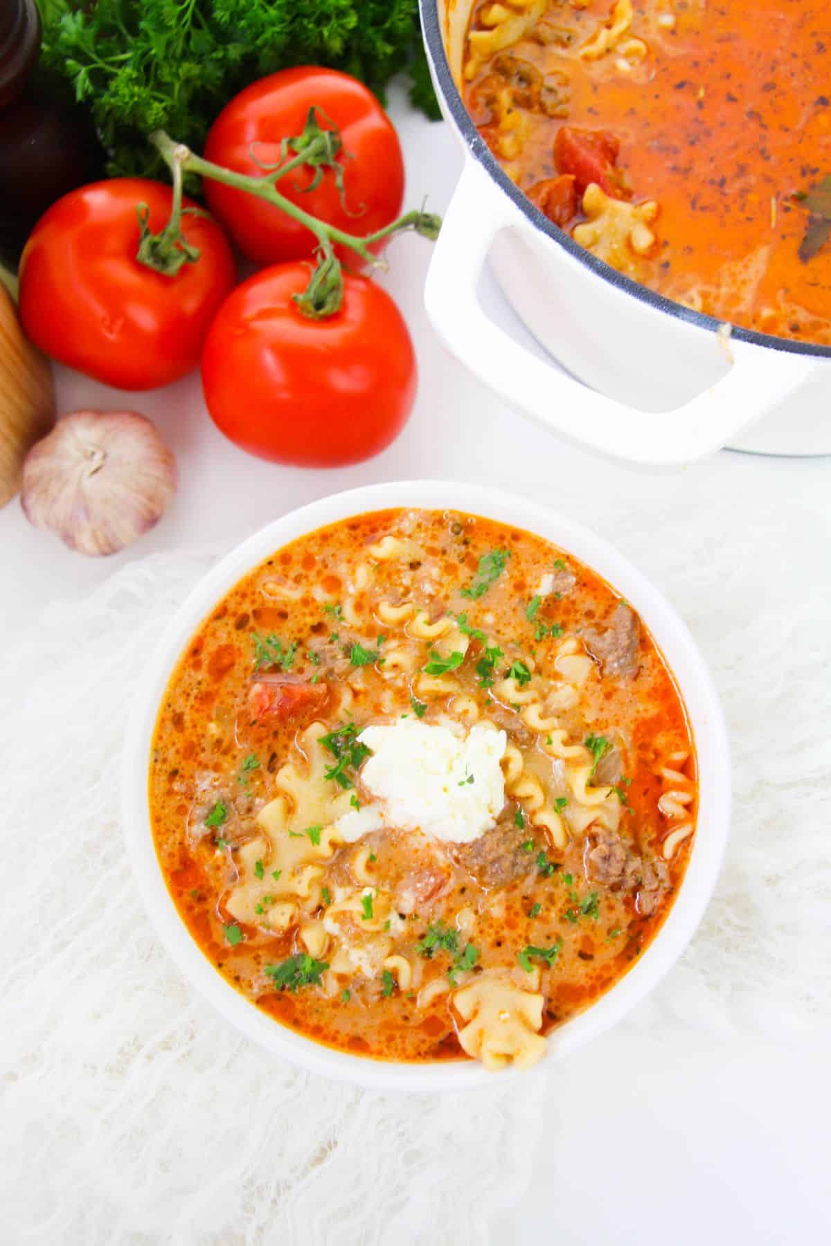 One Pot Lasagna Soup in a serving bowl with a pot, tomatoes, onion and parsley on the side.