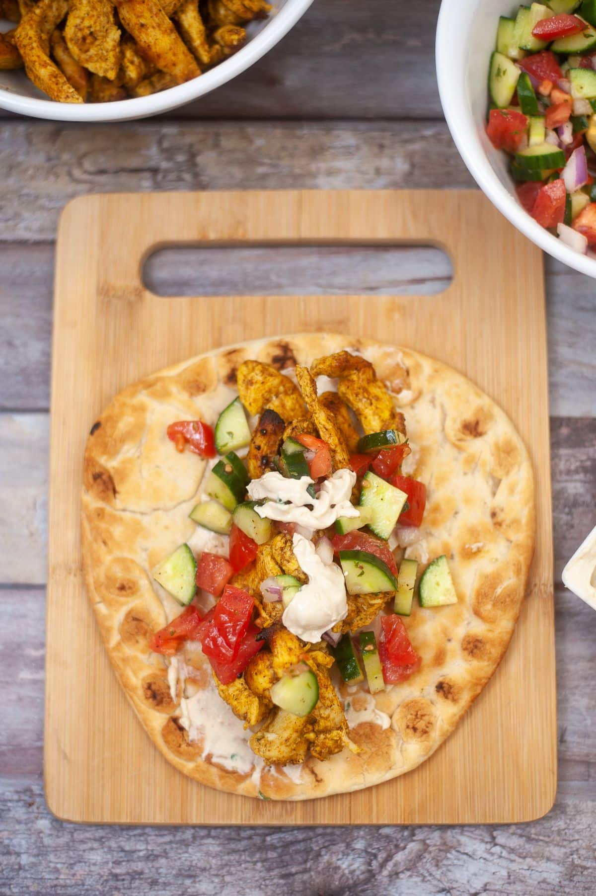 pita bread, lemon tahini sauce, chicken slices, and salad arranged on a wooden chopping board.