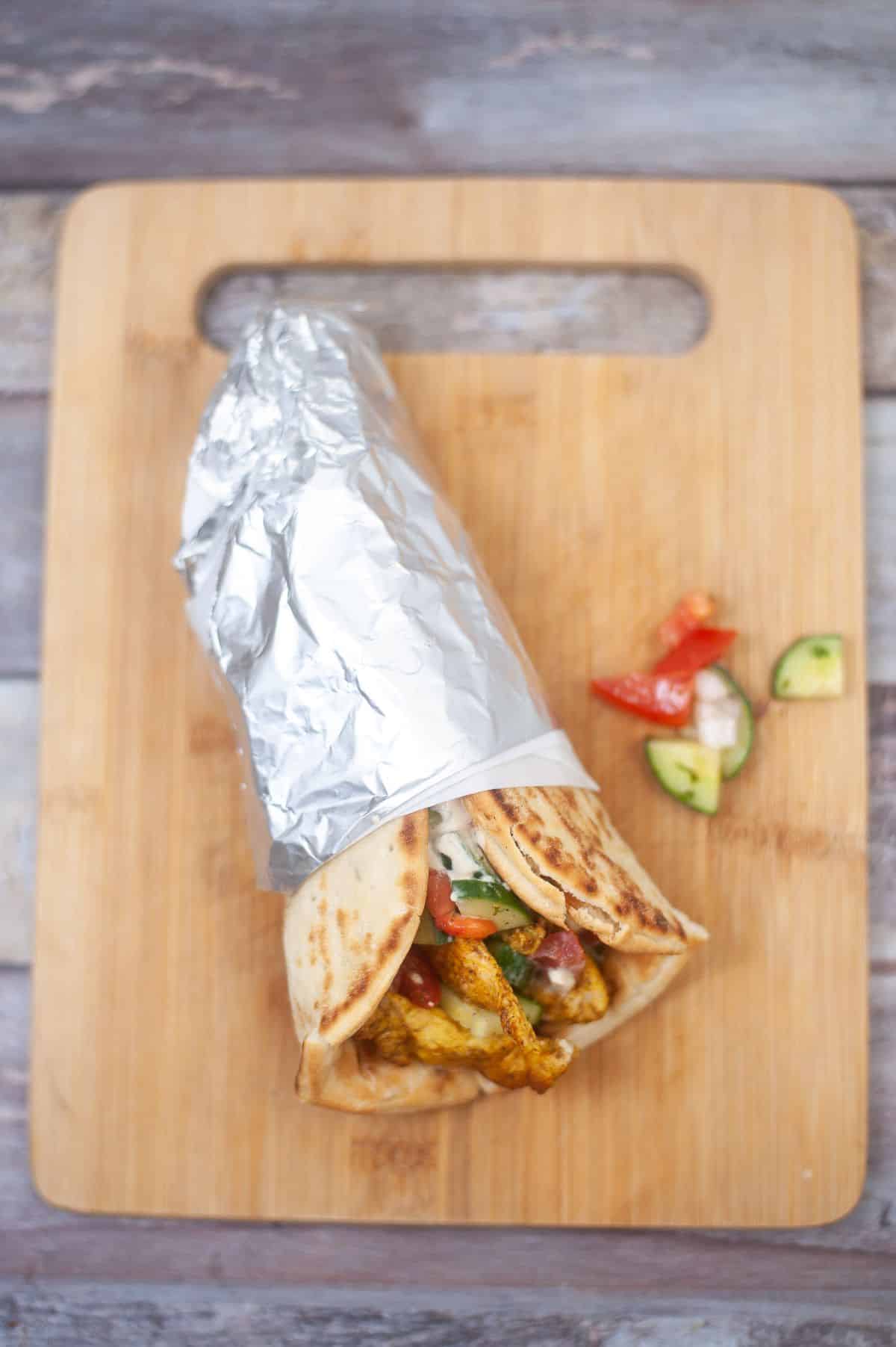 Chicken shawarma wrapped in foil arranged on a wooden chopping board.