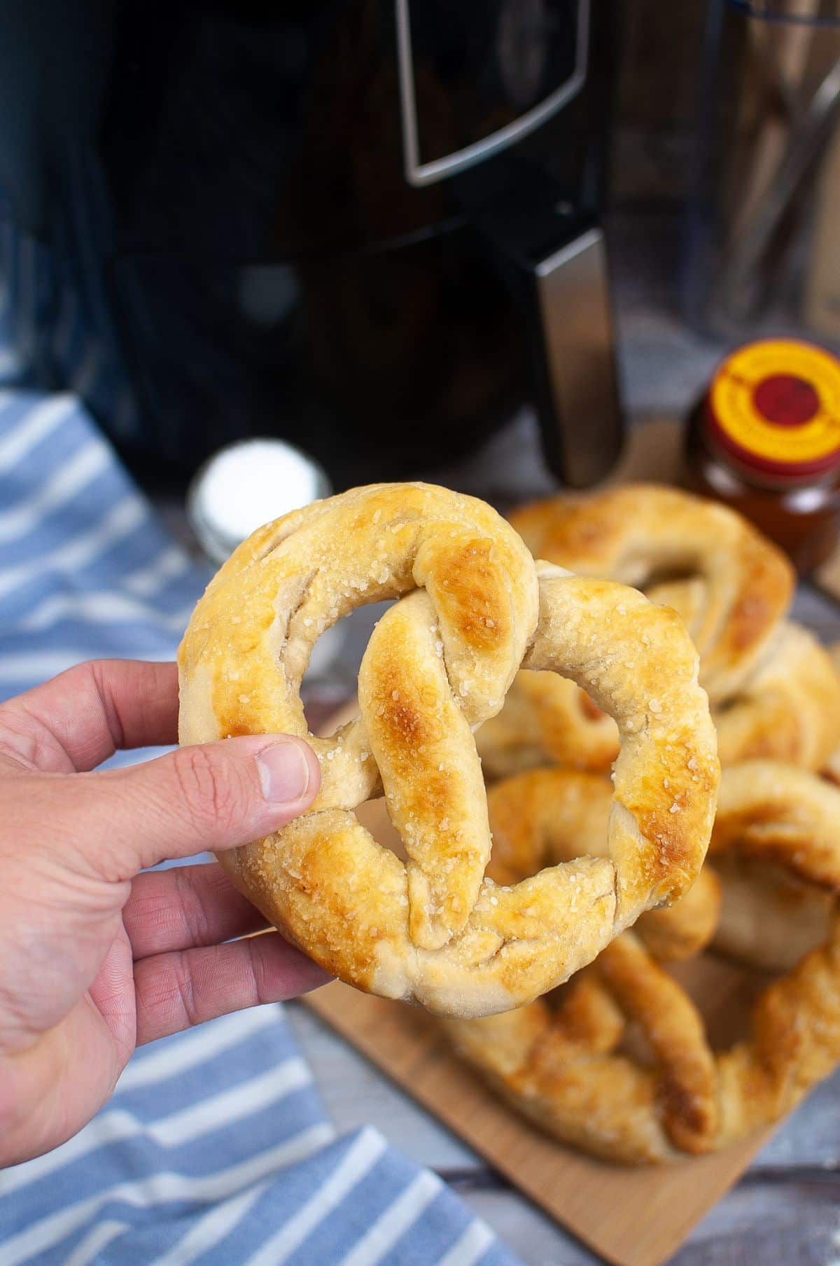 A hand holding pretzel above more pretzels with Air Fryer, salt and sauce on the side. 