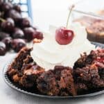 A plate of Chocolate Cherry Cobbler topped with whipped cream and a cherry, with fresh cherries in the background.