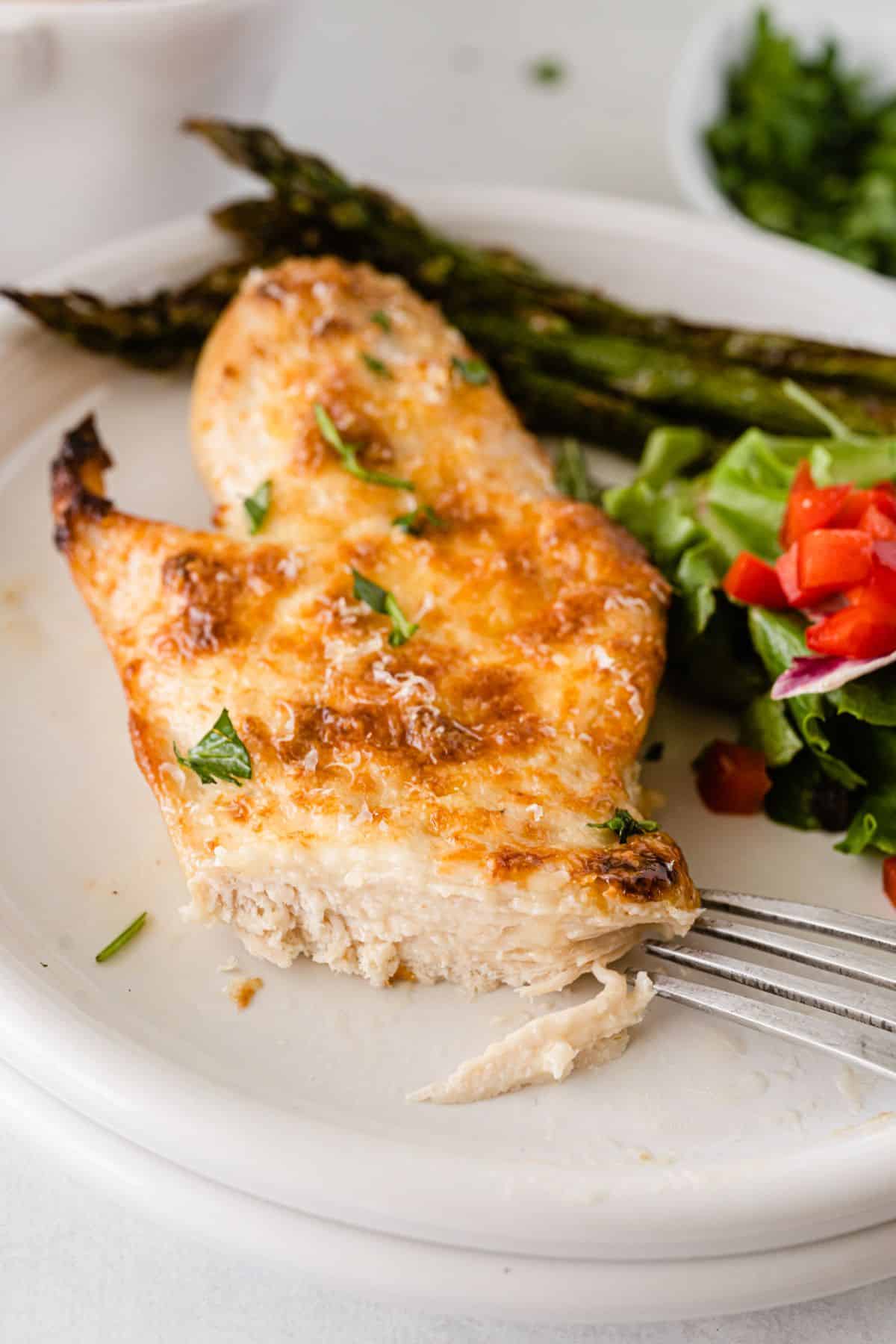 a closeup of Mayo Parmesan Chicken on a serving plate with a fork, asparagus and salad on the side.