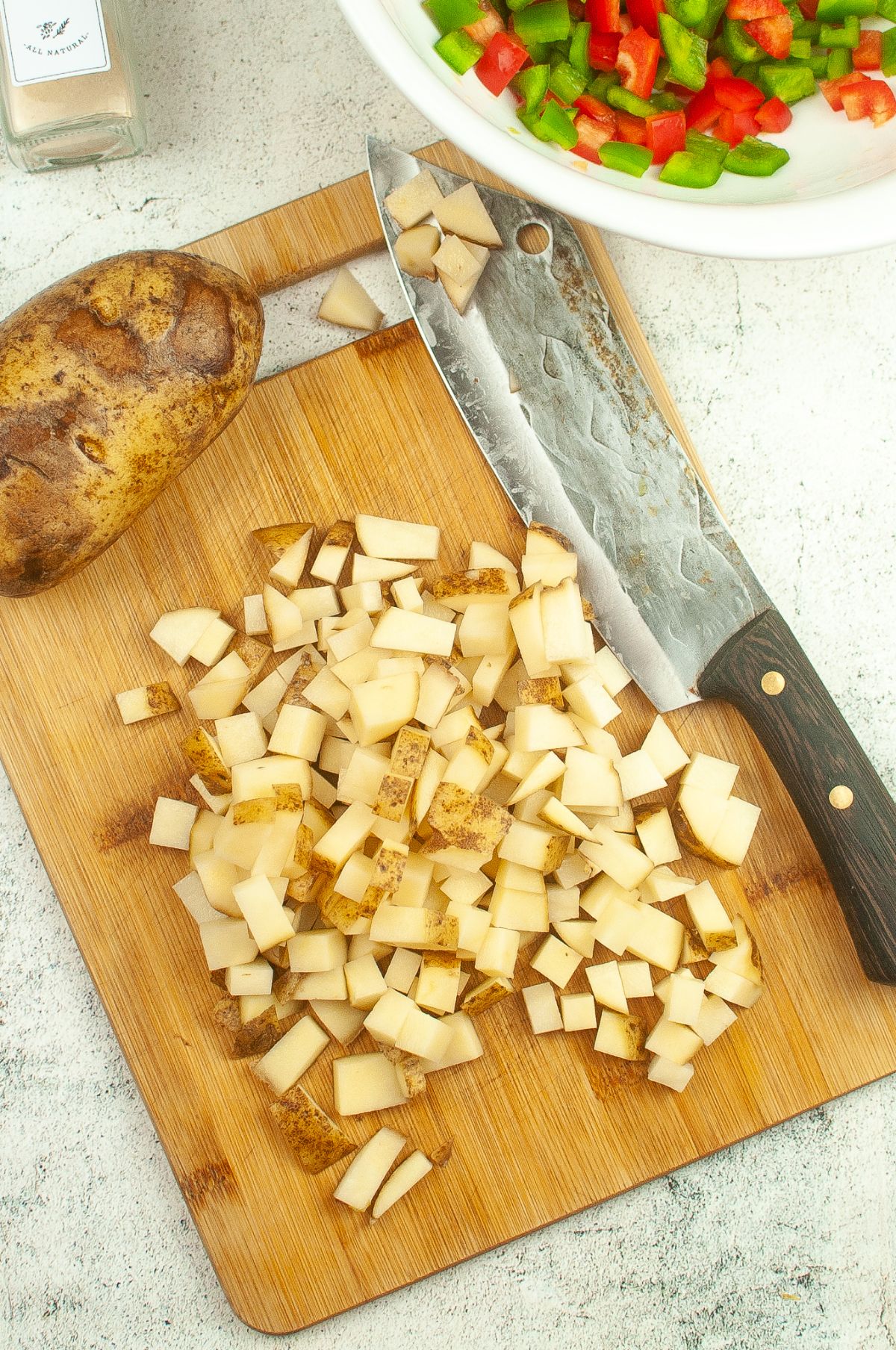 Chopped potatoes on a wooden chopping board with knife and bowl of chopped peppers on the side.