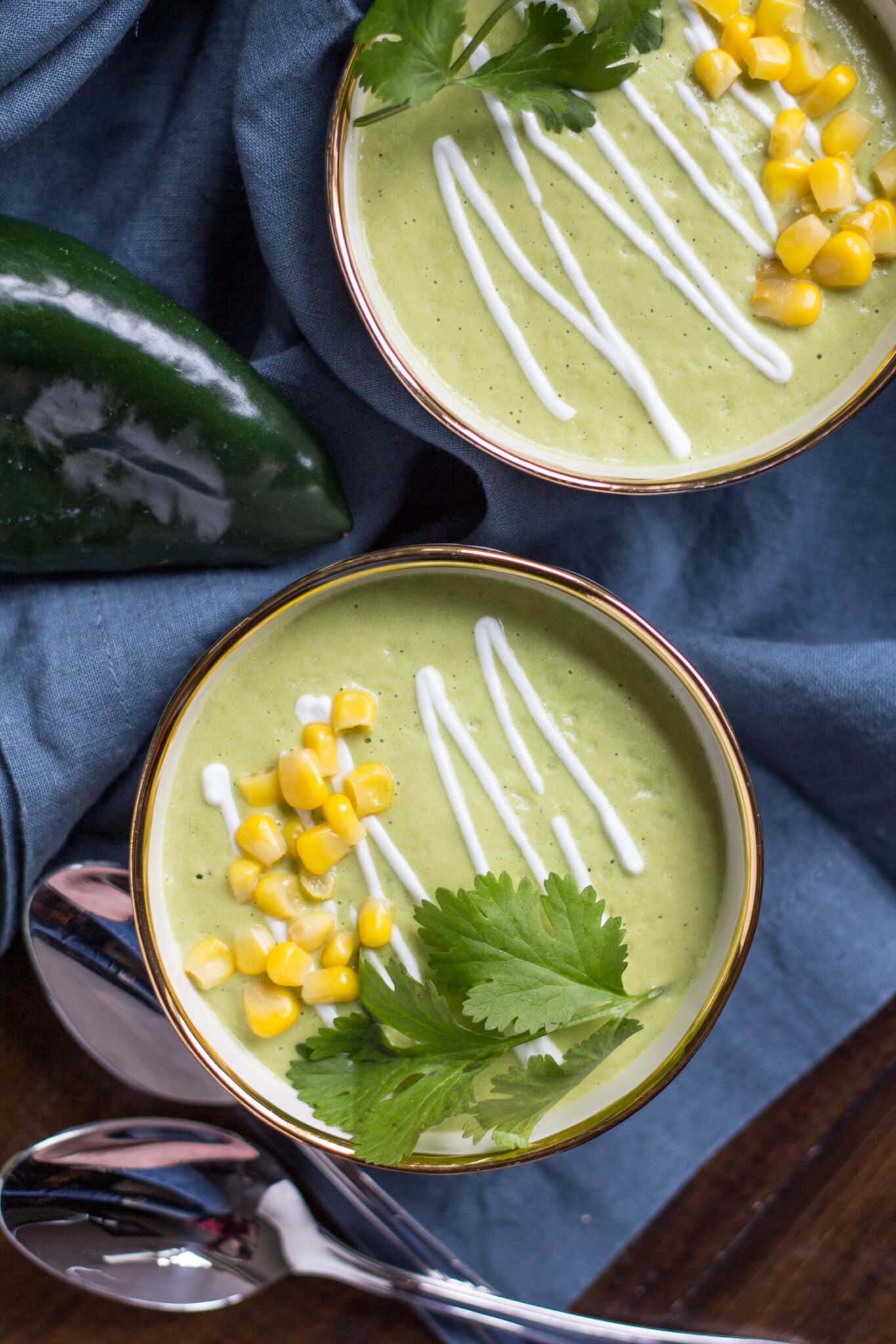 2 bowls of Avocado Poblano Soup on a blue cloth, garnished with cilantro, sour cream, and corn.