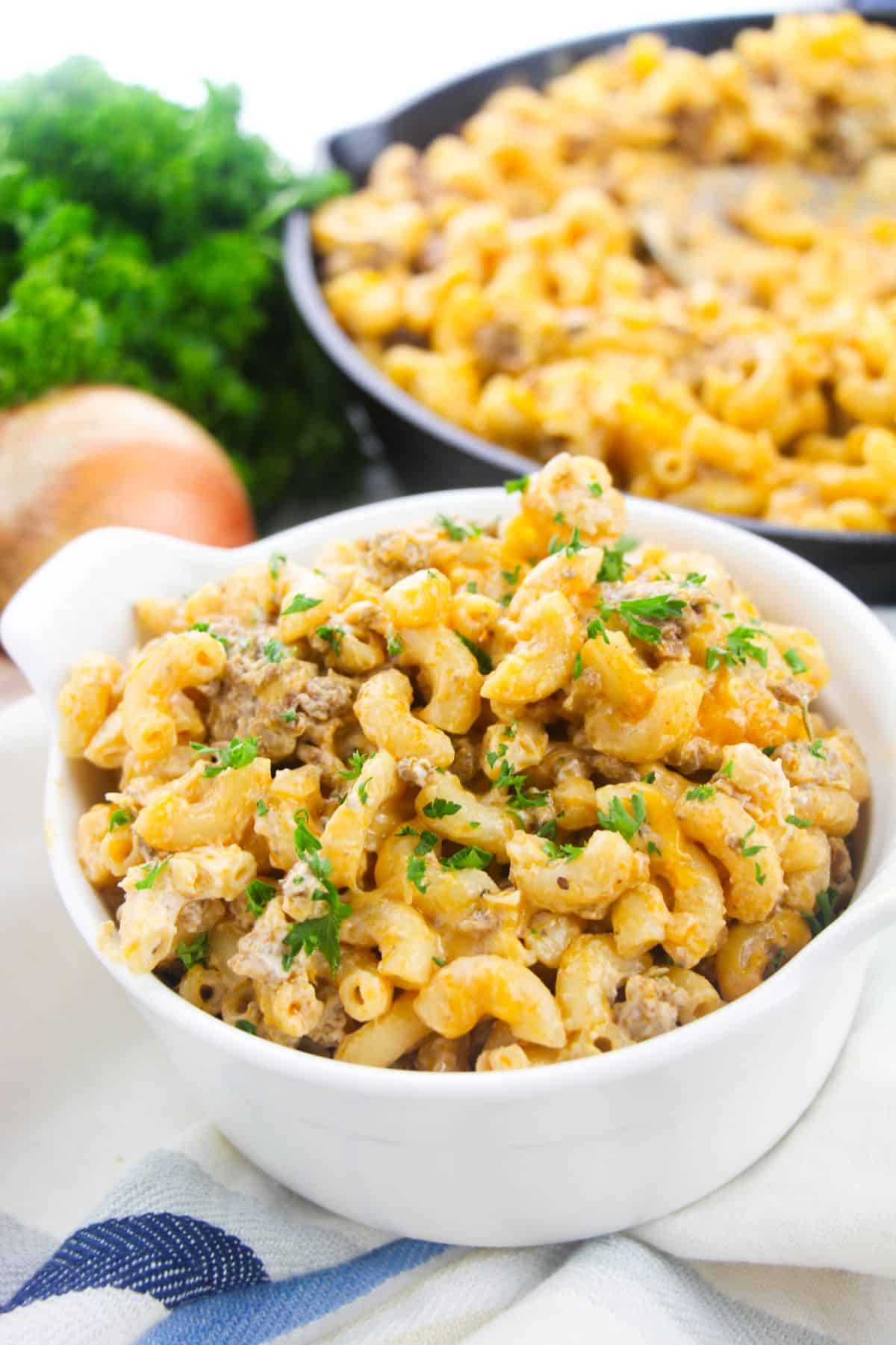 Cheeseburger Macaroni in a bowl, with more Cheeseburger Macaroni in a large skillet , onion and parsley on the side.