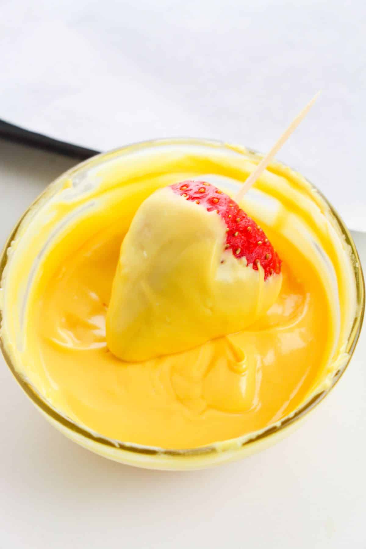 strawberry with a toothpick is being dipped into the yellow melted candy in a small bowl.
