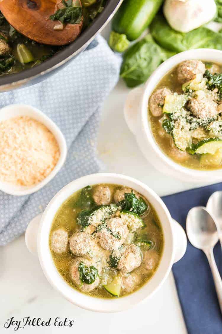 2 bowls of Italian Wedding Soup, garnished with parmesan cheese. Zucchini, spinach,  parmesan cheese, spoons on the side.