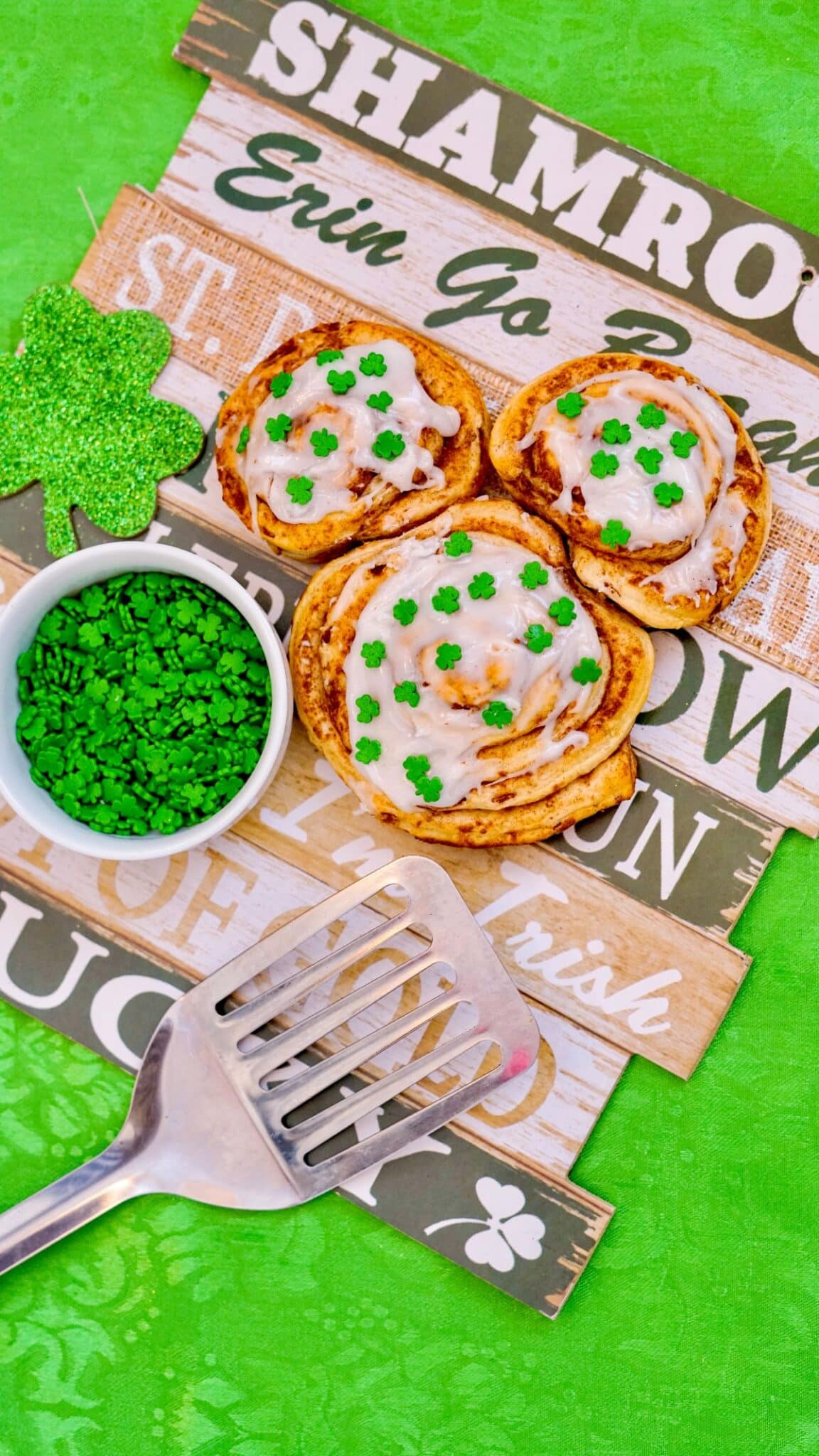 Mickey Cinnamon Rolls on a printed wooden board. St. Patrick's Day sprinkles and spatula on the side.