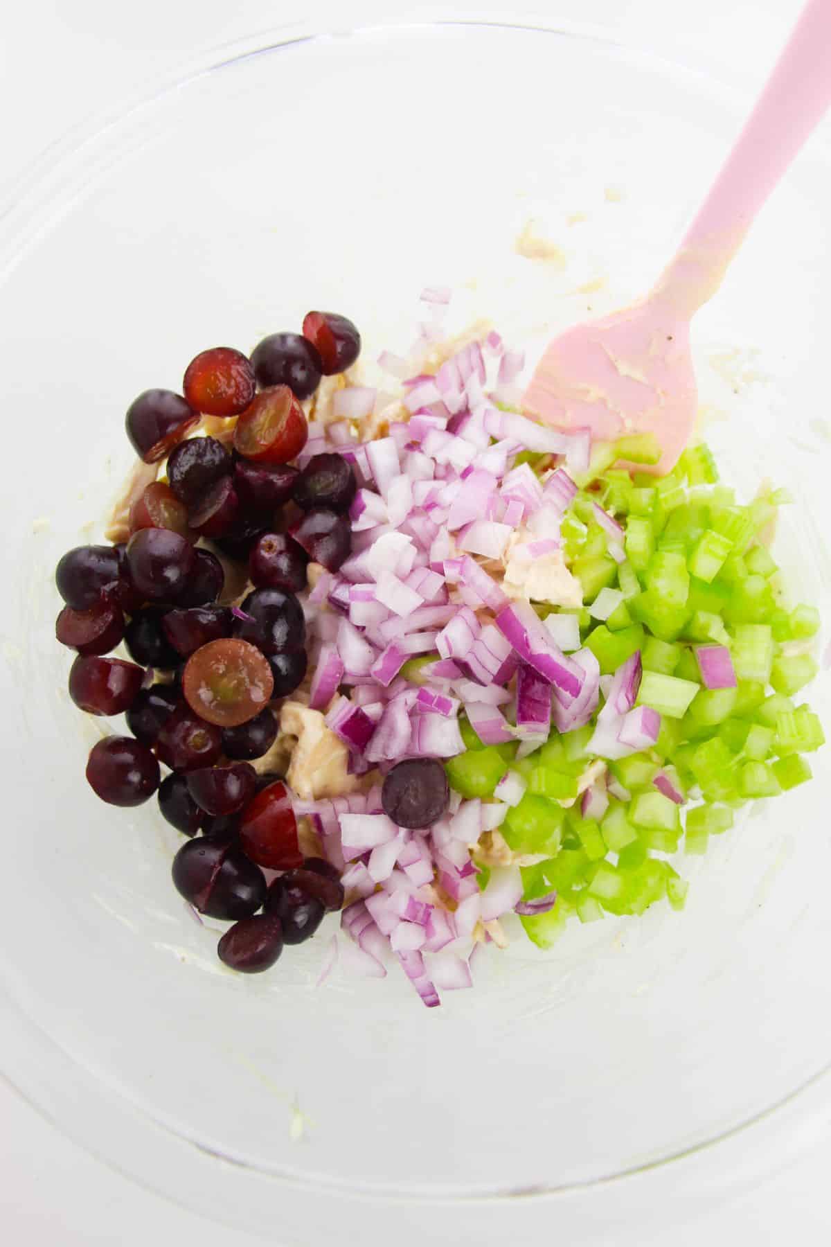 celery, onion, and grapes are added to the mixture in a large bowl with pink spatula.