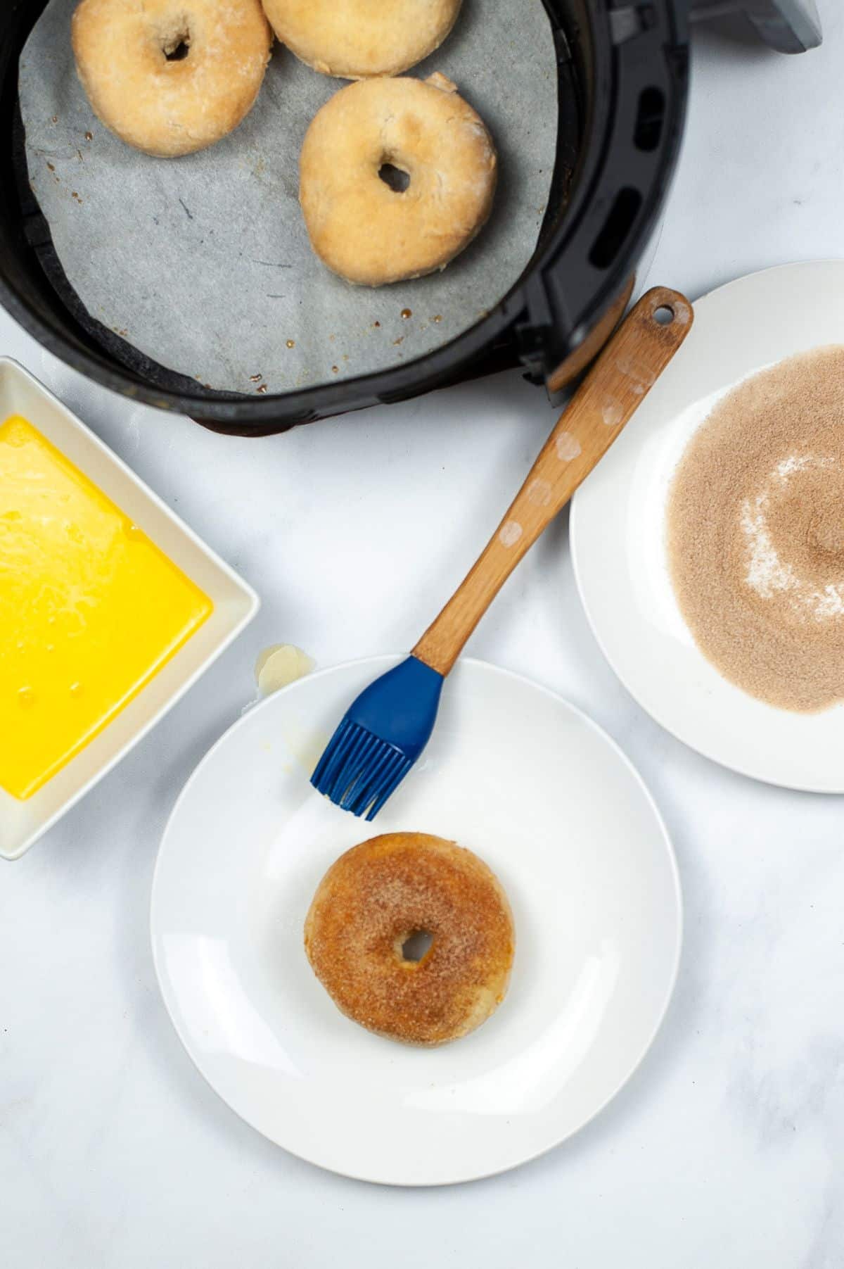 a coated donut on a white plate next to a blue pastry brush, next to a bowl of melted butter, plate of cinnamon sugar and donuts in an air fryer